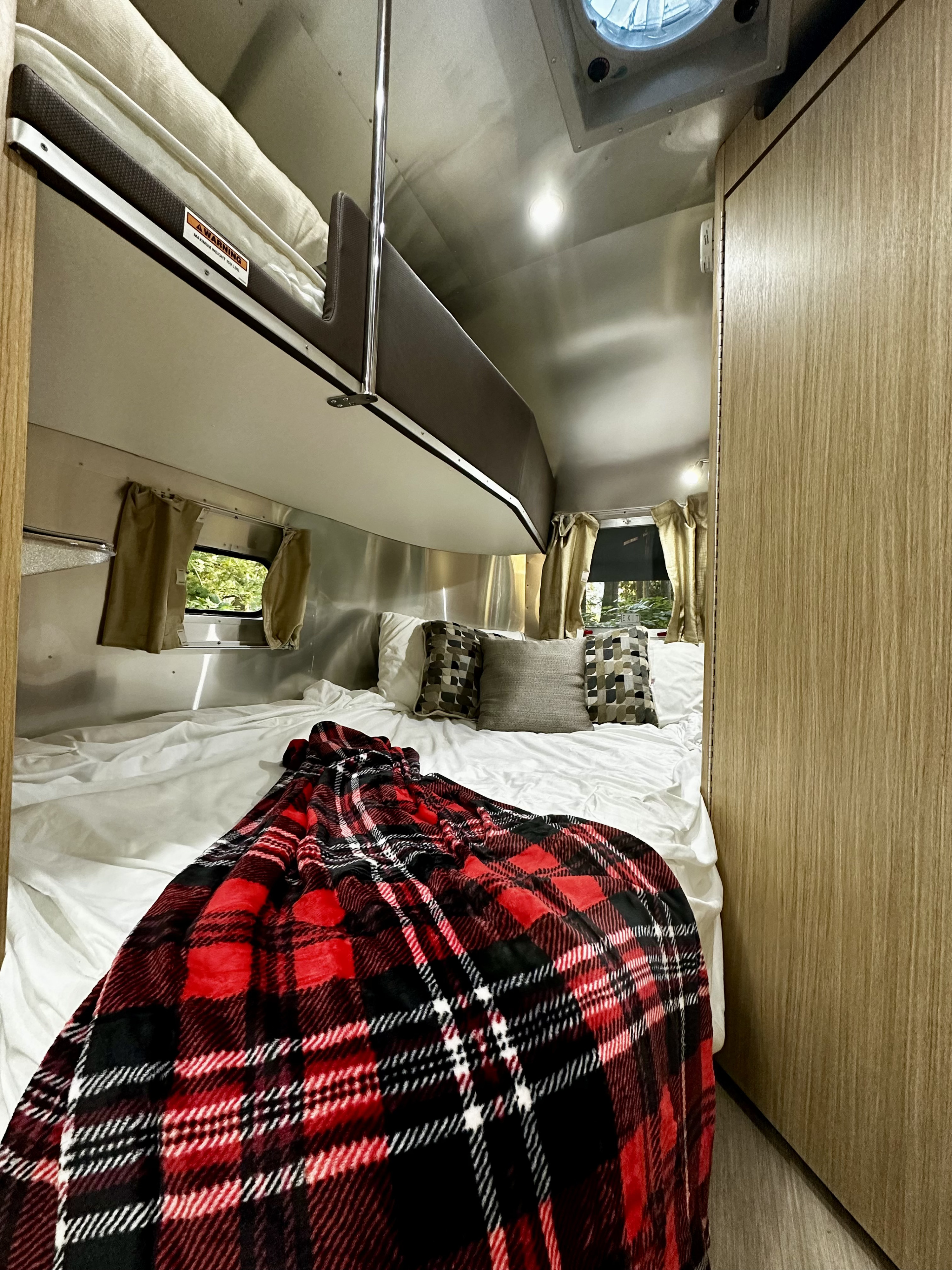 Cozy RV rental interior: main bed with red plaid blanket, overhead bunk, and bright aluminum walls for a comfy trip.. Airstream Flying Cloud 2019