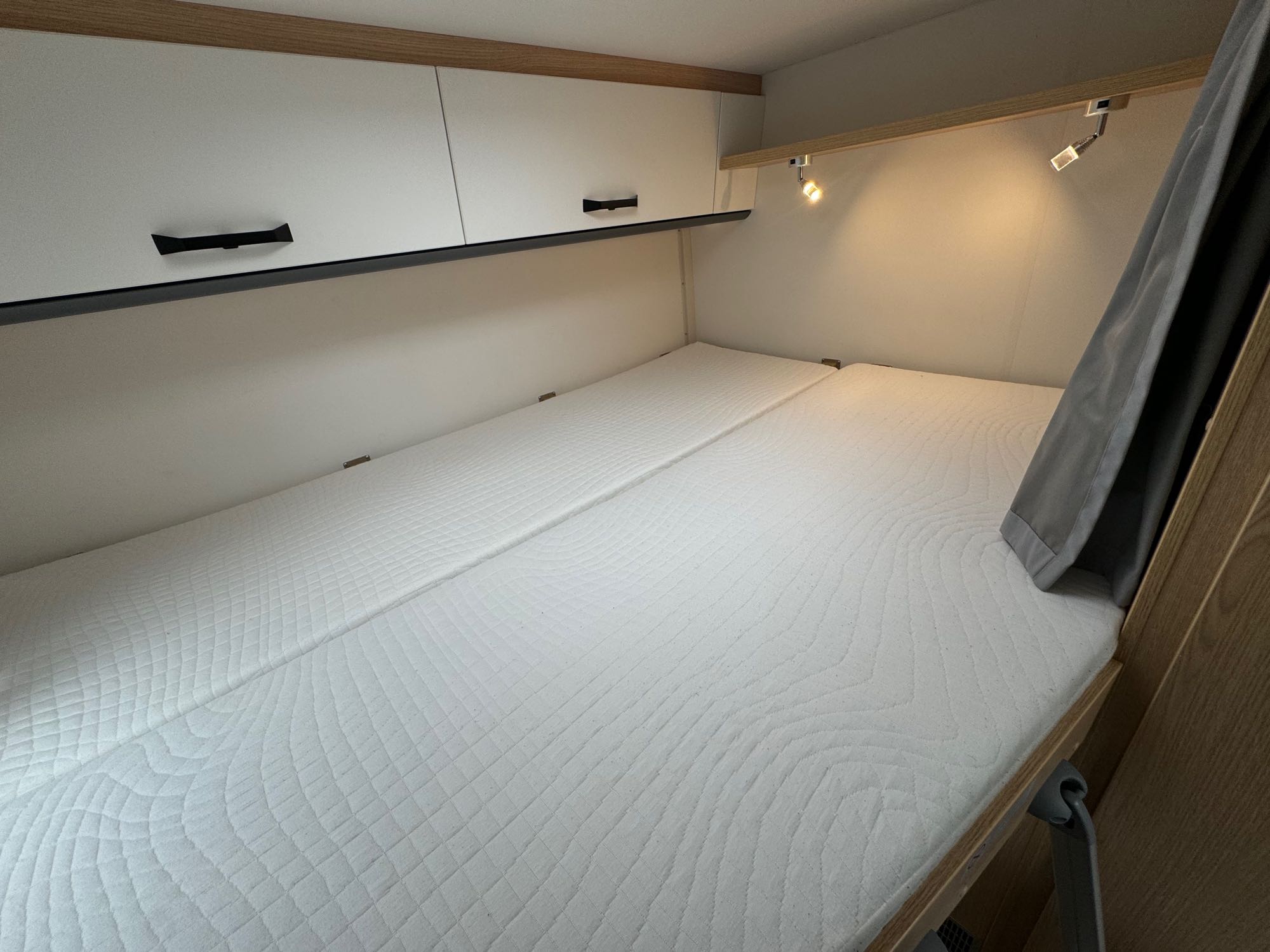 Cozy RV rental sleeping area featuring two white quilted mattresses, overhead storage cabinets, and functional reading lights.. Sunlight V60 2025