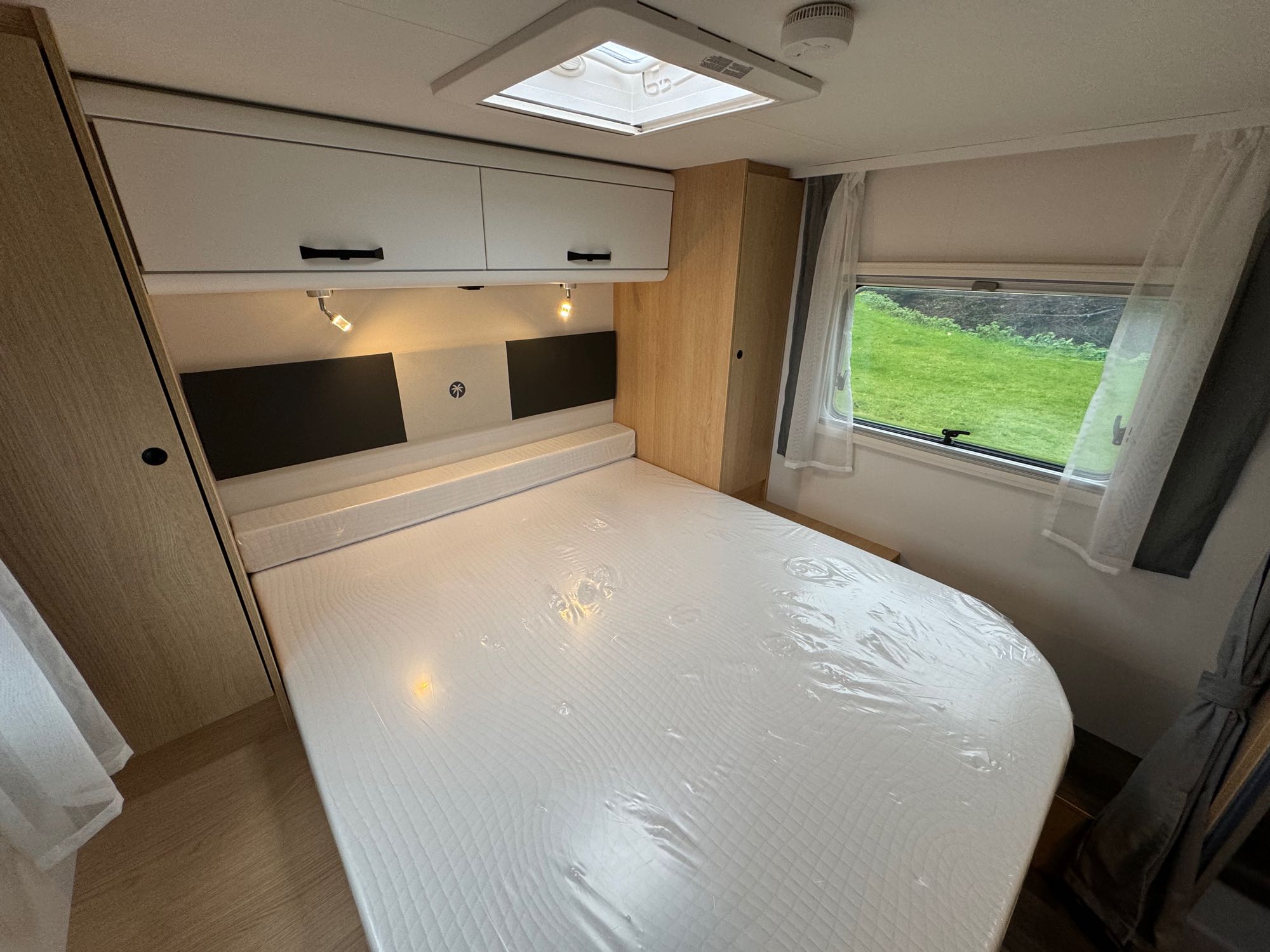 Inside an RV rental: a bright bedroom features a new, plastic-covered mattress, overhead cabinets, and a window overlooking greenery.. Sunlight 2025