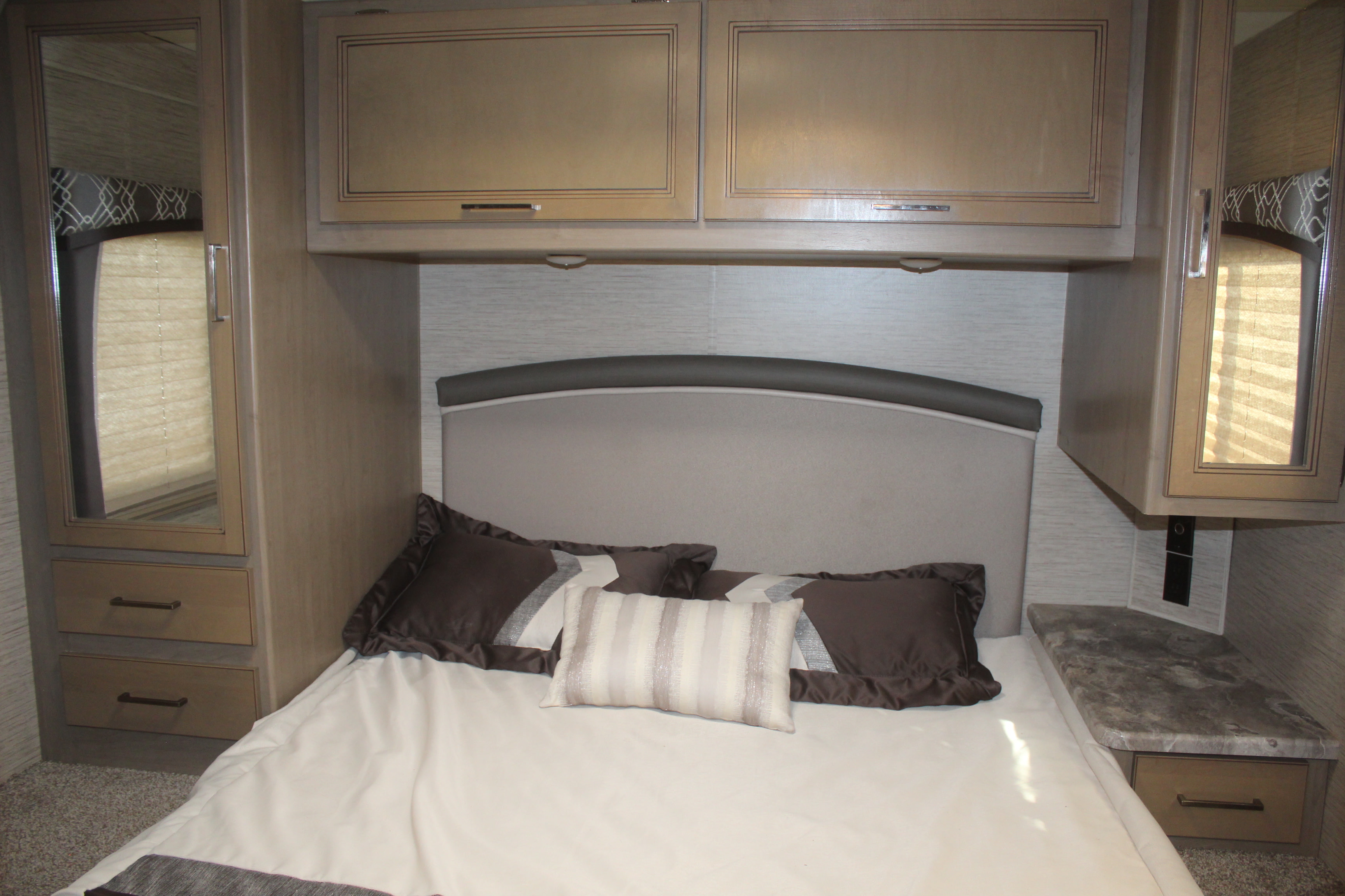 RV rental bedroom with a neatly made bed, padded headboard, and overhead storage. Mirrored cabinet and drawers on left, counter on right.. Thor Chateau 2021
