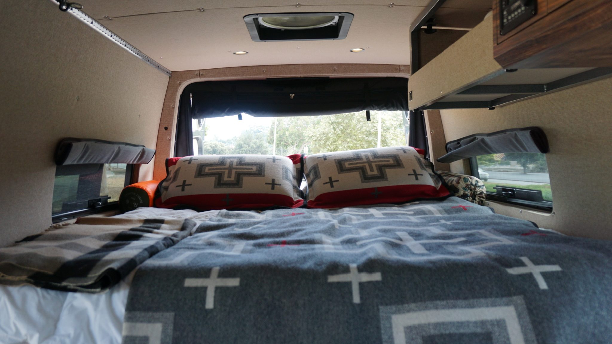 Cozy RV rental interior: a comfy bed with patterned blankets & pillows, featuring large windows & a ceiling vent.. Mercedes-Benz Sprinter 2021