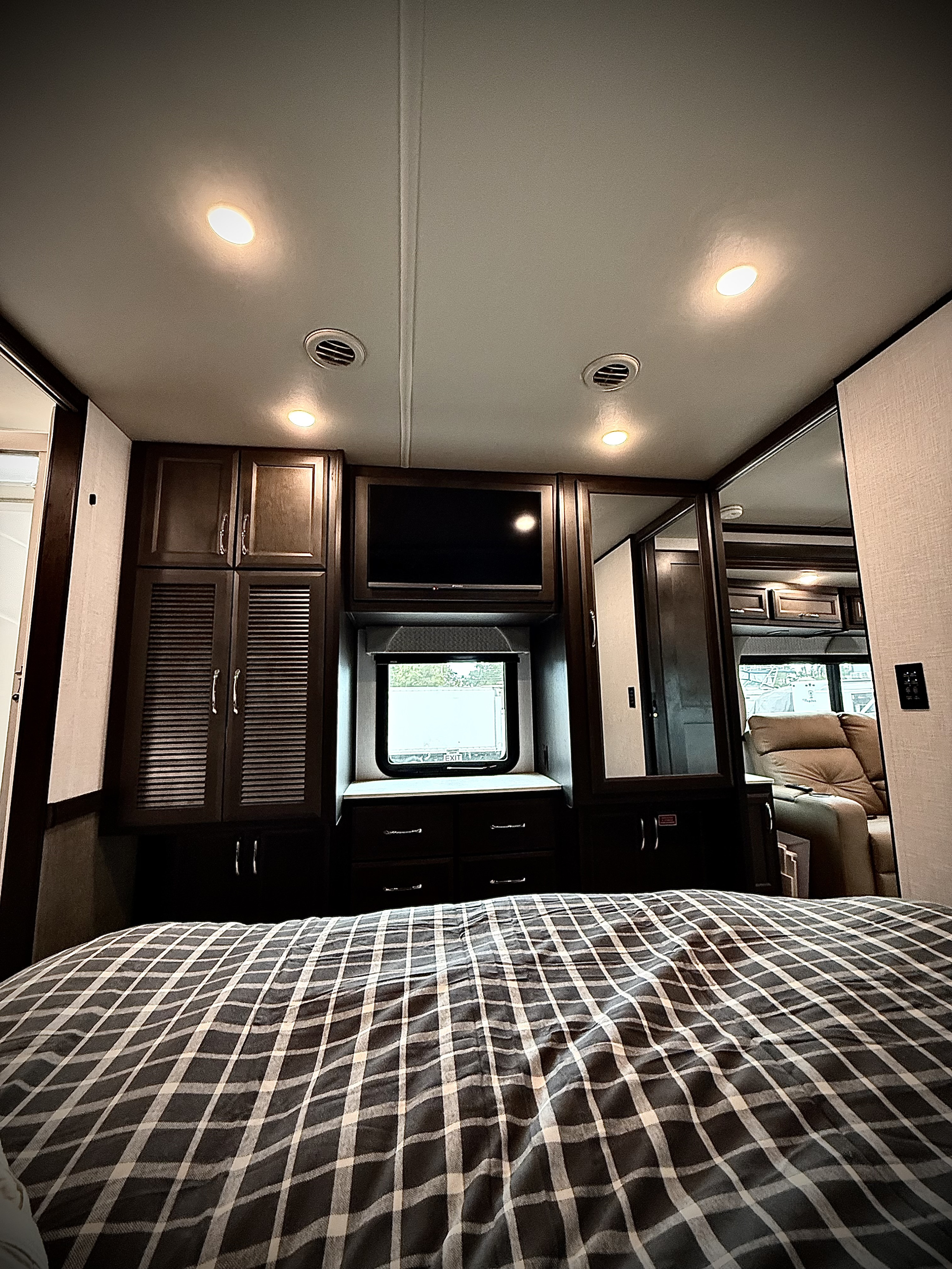Cozy RV rental bedroom: plaid bed, dark wood cabinets, TV, and mirrored closet reflecting the living area.. Bounder Bounder Motorhome 2024