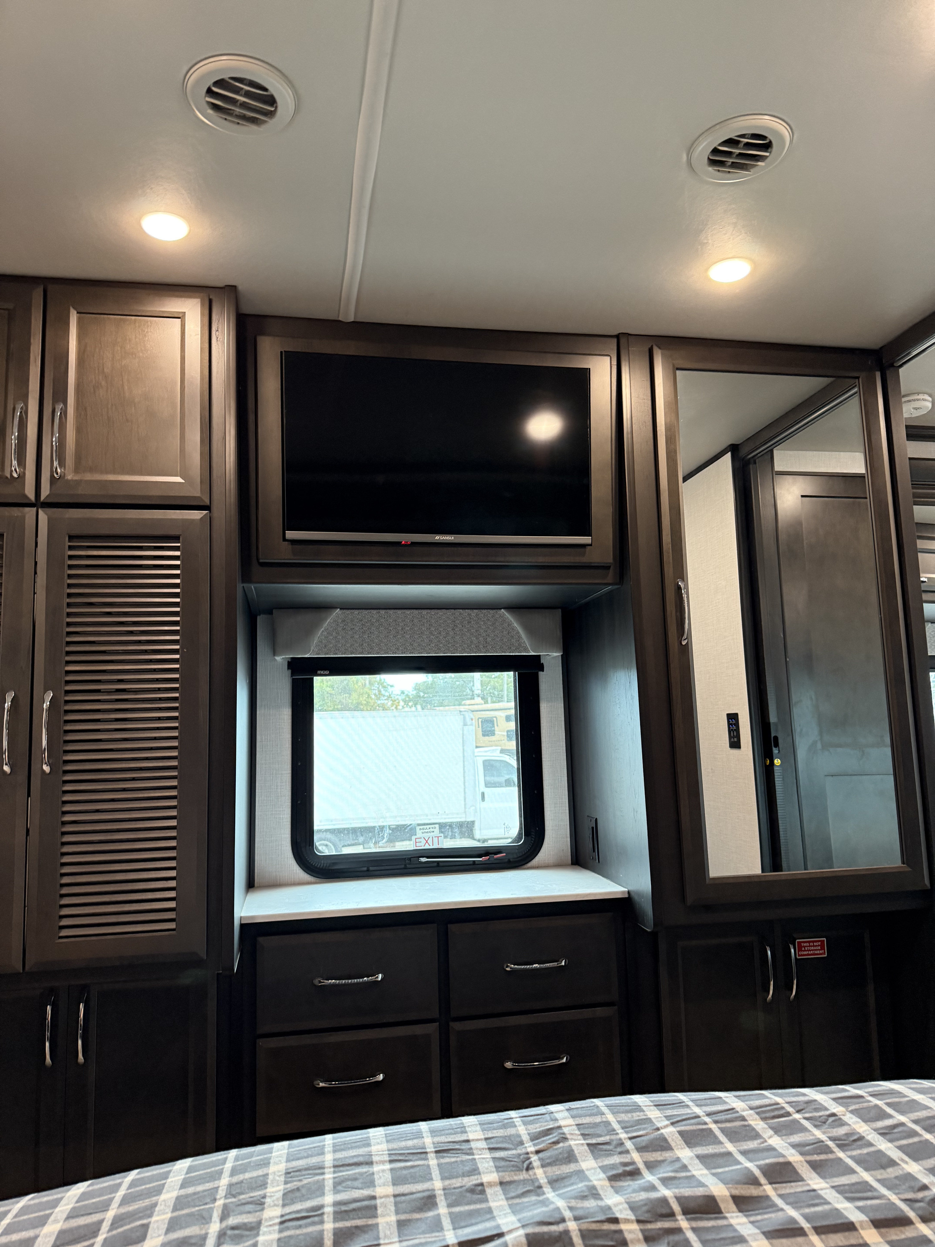 An RV rental bedroom with a mounted TV, dark wood cabinets, a window, drawers, and reflective mirror. A functional and comfortable space.. Bounder Bounder Motorhome 2024