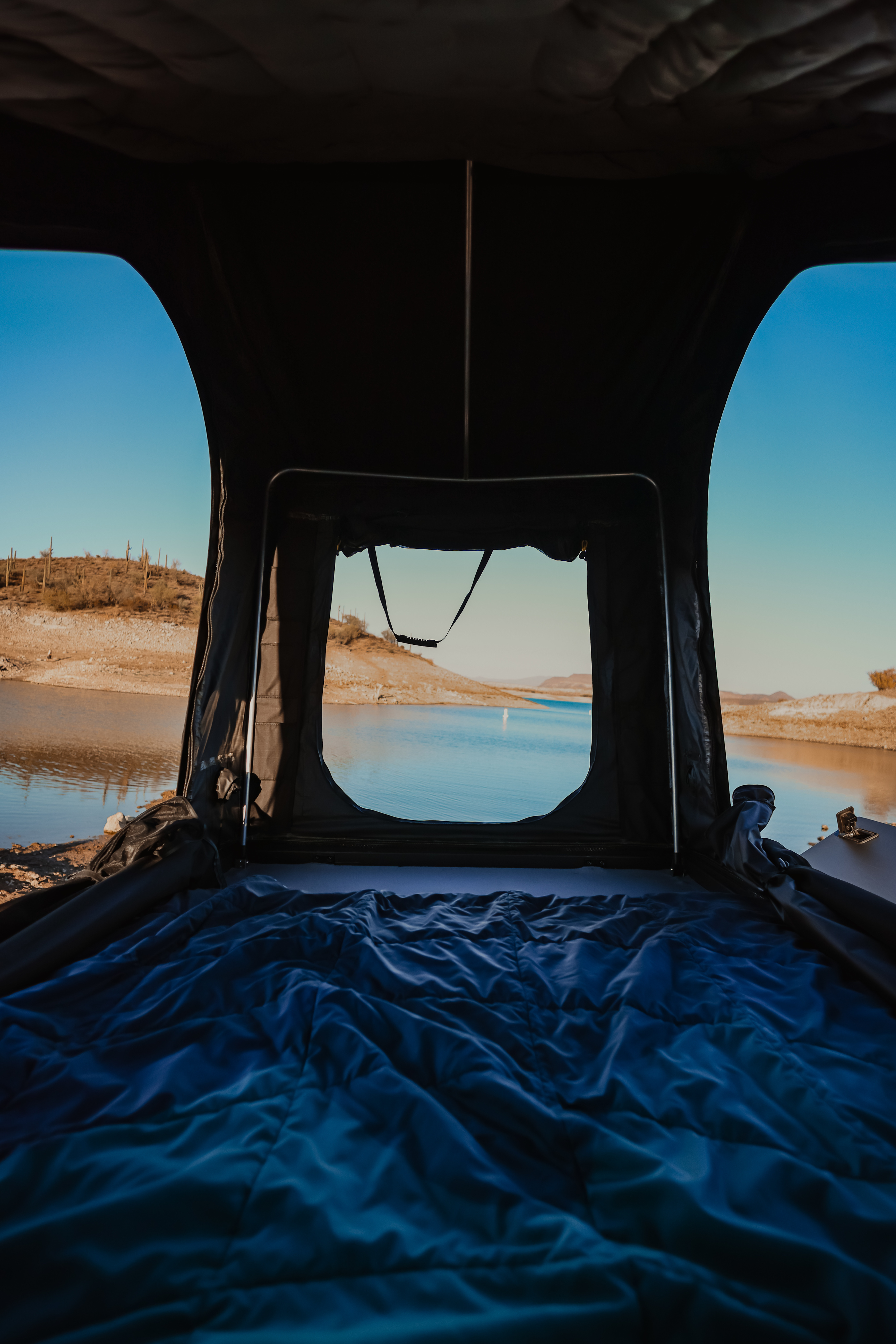 Cozy rv rental rooftop tent with a blue comforter, offering serene views of a desert lake and distant cacti.. Jeep Gladiator 2024