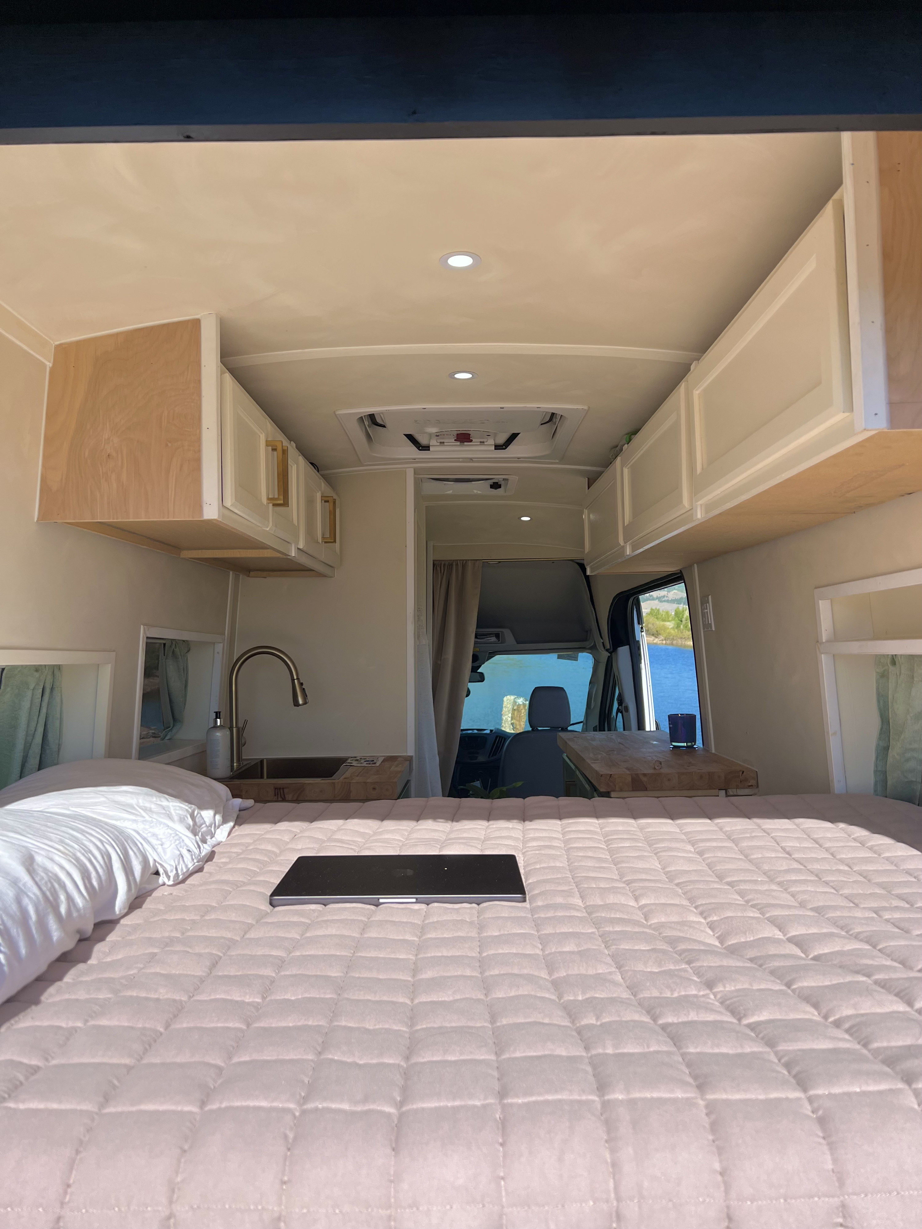 Cozy RV rental interior: foreground bed with laptop, midground kitchen sink, and a beautiful lake view beyond the cab.. Ford Transit 2016