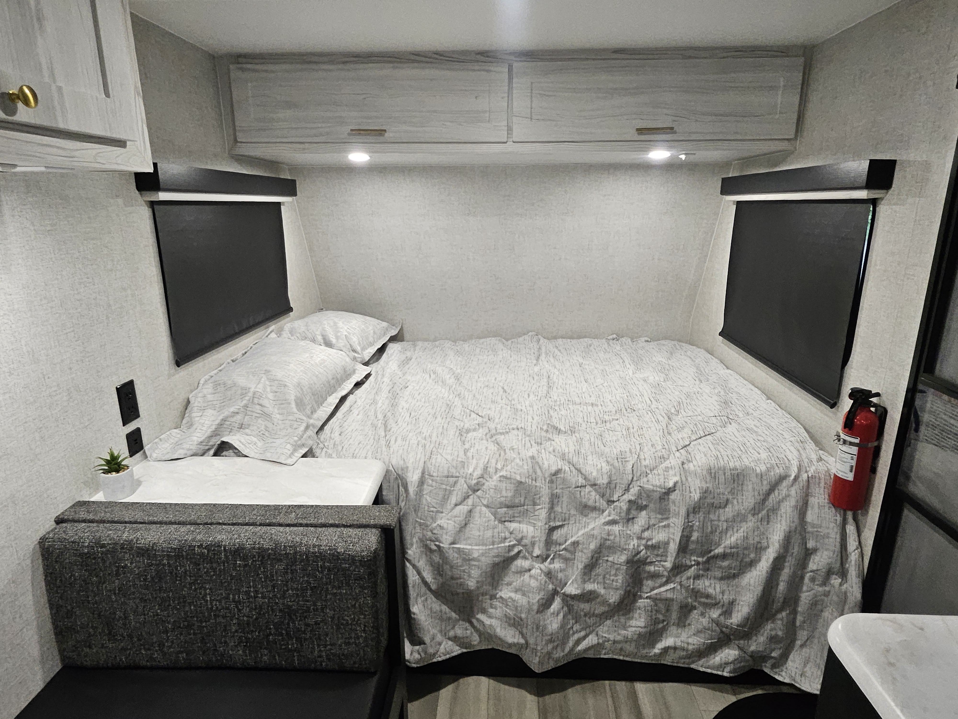 RV rental bedroom view: a neatly made bed with light bedding, overhead cabinets, dark window shades, and a fire extinguisher.. Eclipse Milan 16MBH 2024