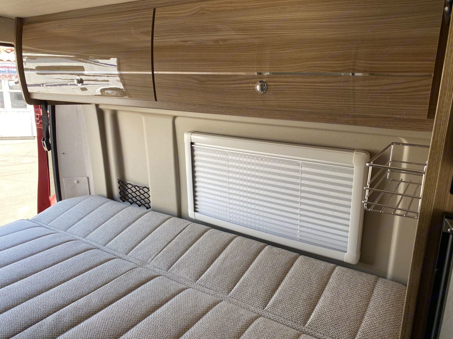 Cozy RV rental: gray quilted bed, glossy wood overhead cabinets, and a window with blinds for comfortable travels.. Winnebago Travato 2020