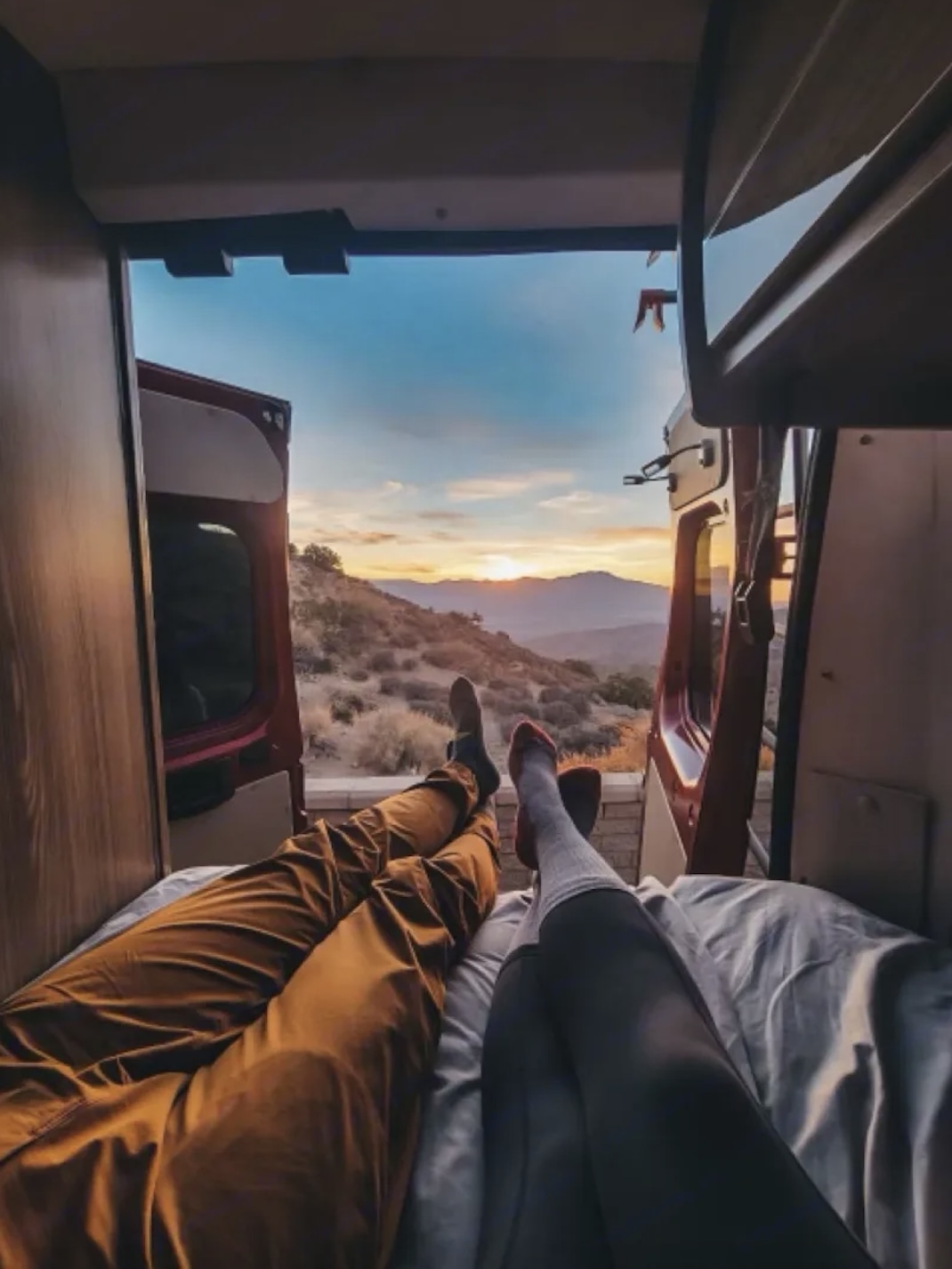 View from inside an RV rental: Two sets of legs relaxed, facing a mountain sunset. Comfort and adventure await.. Winnebago Travato 2020