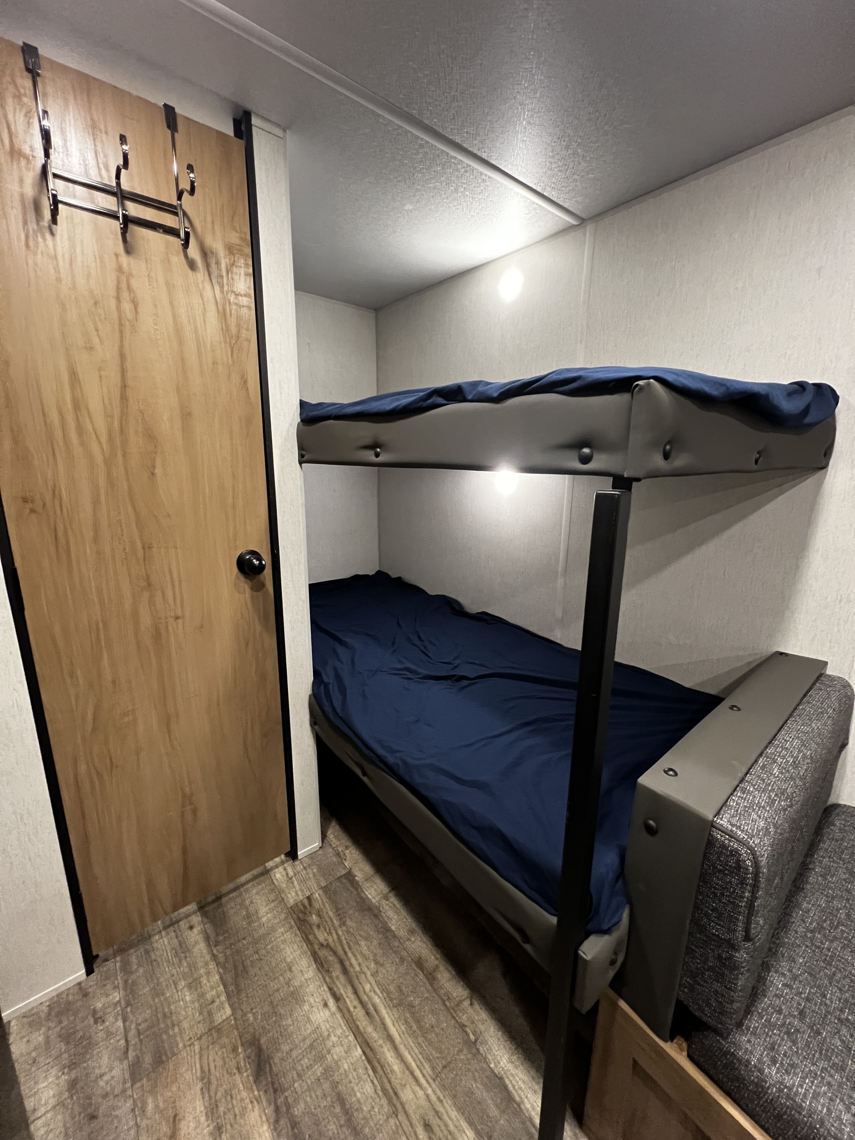 RV rental interior shows compact bunk beds with blue sheets, built-in lights, and an over-door hook rack on left.. Forest River Cherokee Wolf Den 16EV 2024