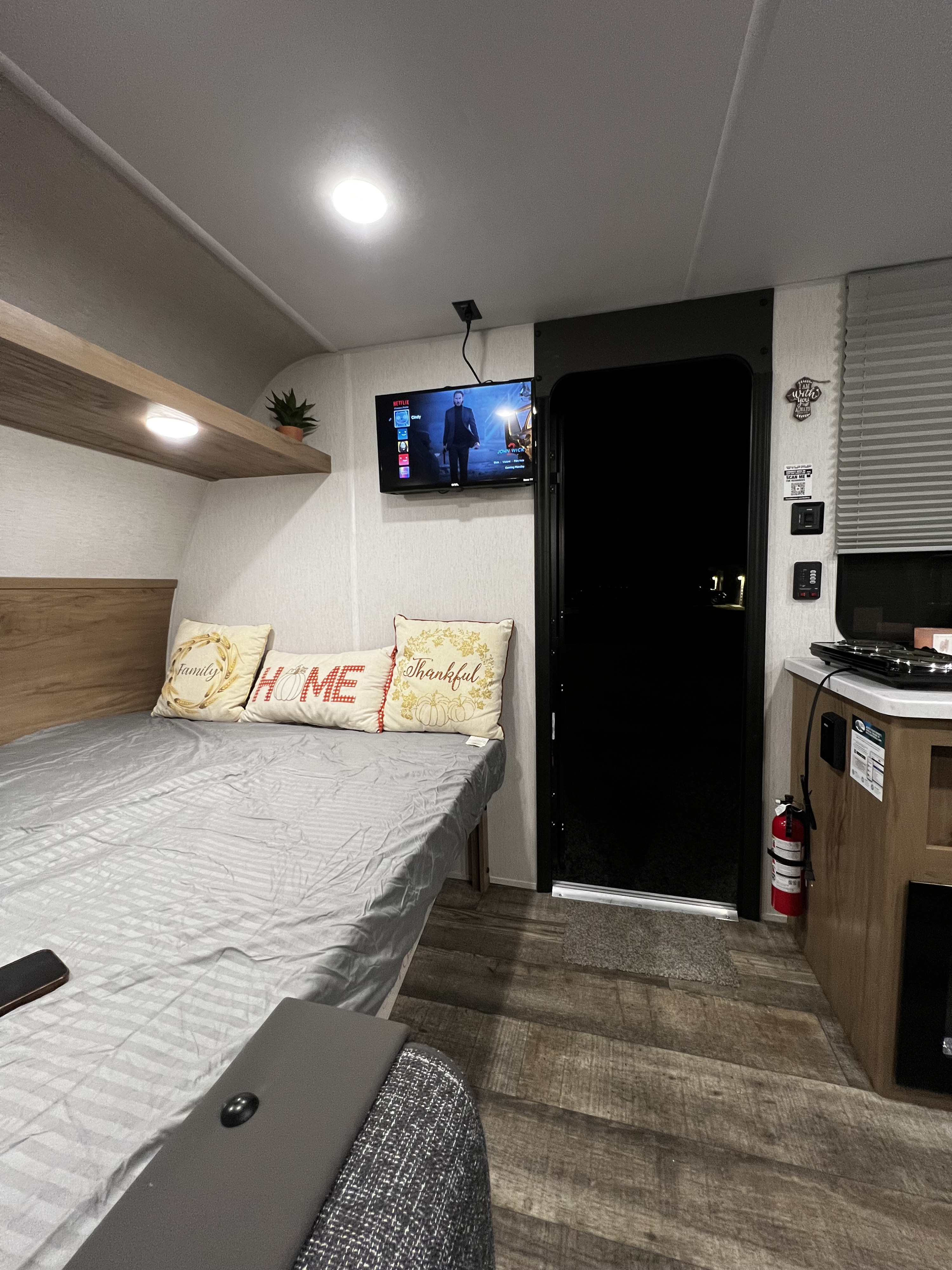Inside a cozy RV rental: a bed with decorative pillows, TV showing John Wick, small kitchen area, and a fire extinguisher.. Forest River Cherokee Wolf Den 16EV 2024