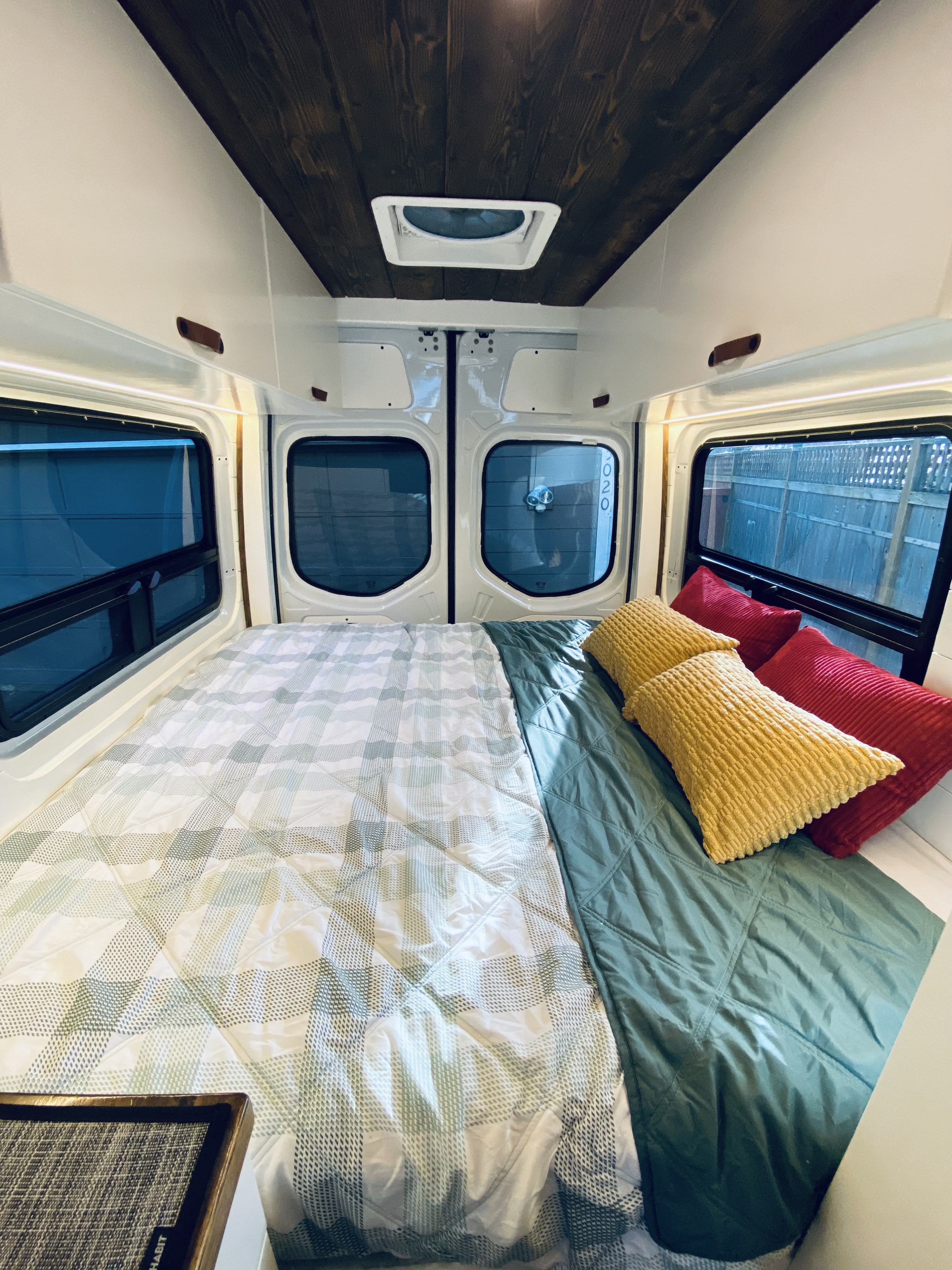 Inside a cozy RV rental: a comfortable bed with plaid/teal bedding, yellow/red pillows, wooden ceiling, and large windows.. Mercedes Benz Mercedes Sprinter 2500 2023