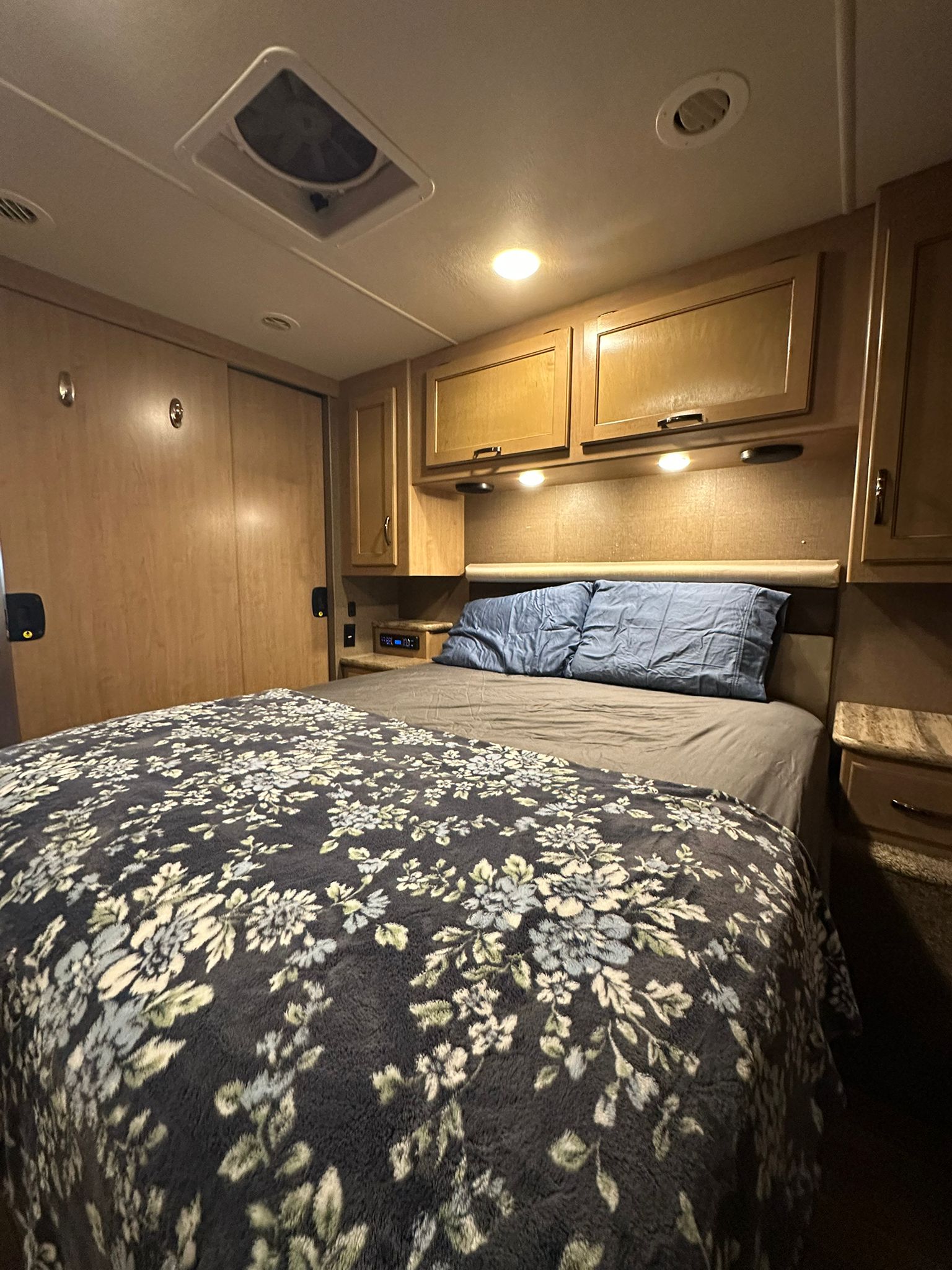 Cozy RV rental bedroom interior with a floral blanket, wood cabinetry, and soft lighting, ideal for travelers.. Thor A.C.E. 2018