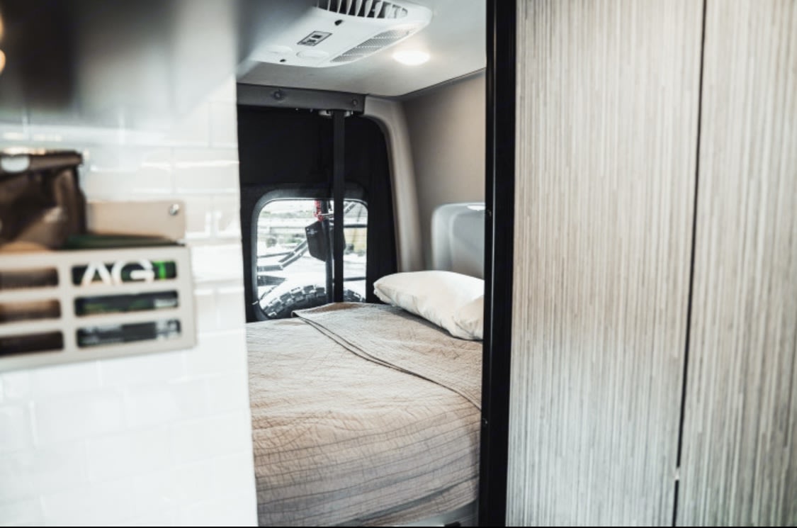 Cozy rv rental interior: a comfortable bed with a quilted blanket and pillow, and an AC unit in a compact camper van.. Winnebago Revel 2022