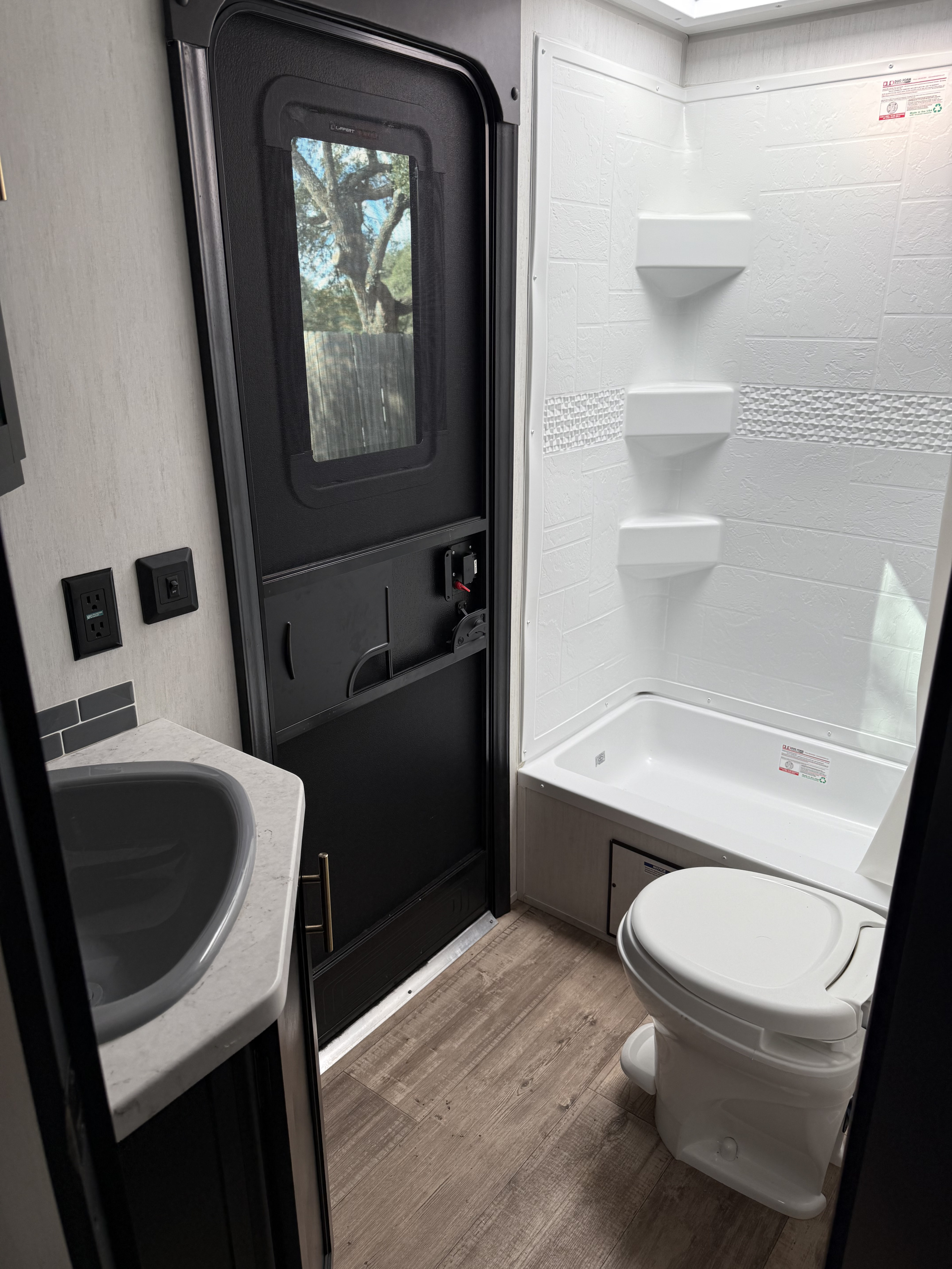 Inside an RV rental bathroom: a modern shower/tub, toilet, and sink. The door reveals a view of trees outside.. Forest River Cherokee Grey Wolf 2022