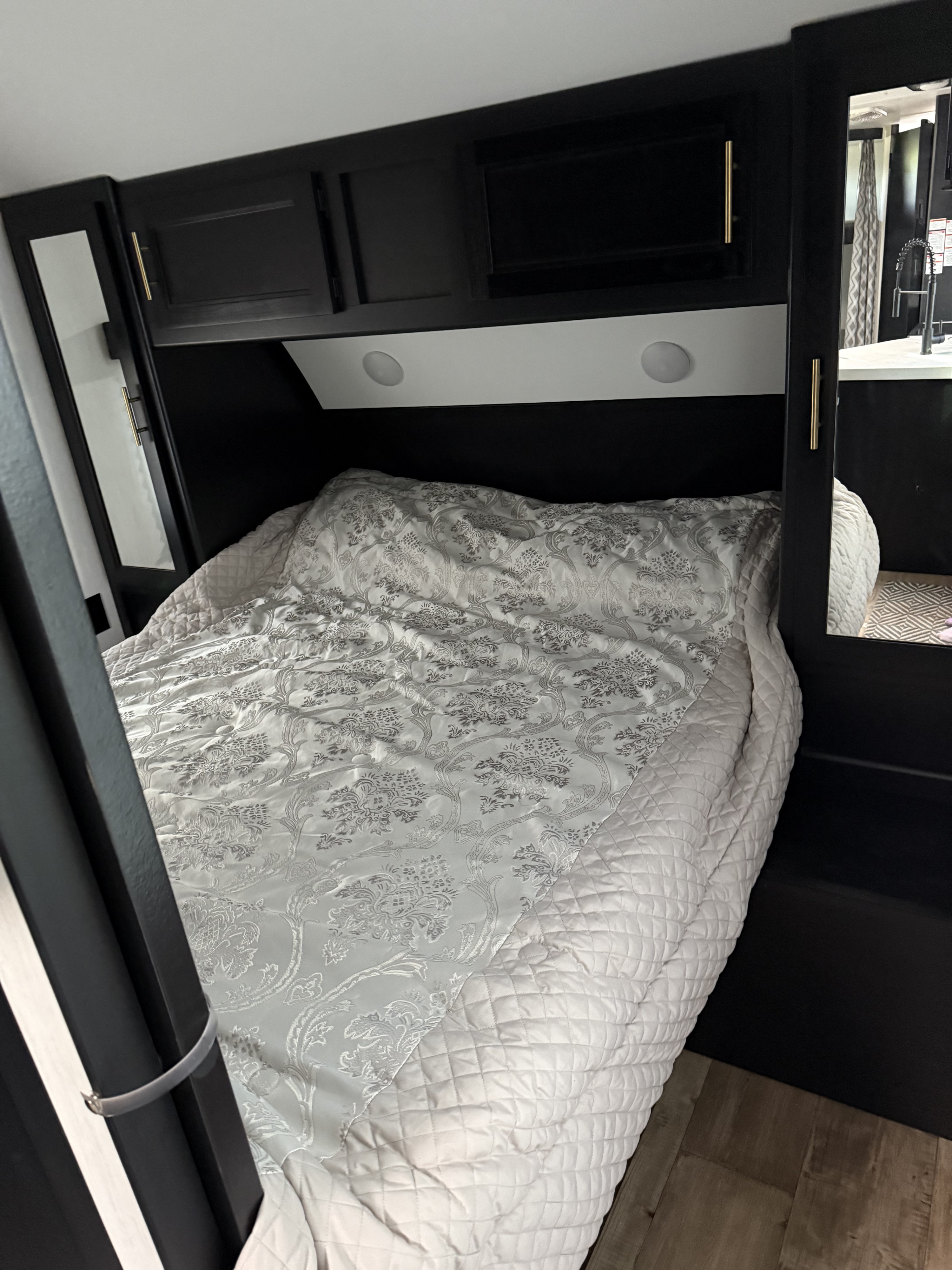 Cozy RV rental bedroom with white patterned bedding, dark cabinets, and mirror reflecting a kitchen area.. Forest River Cherokee Grey Wolf 2022