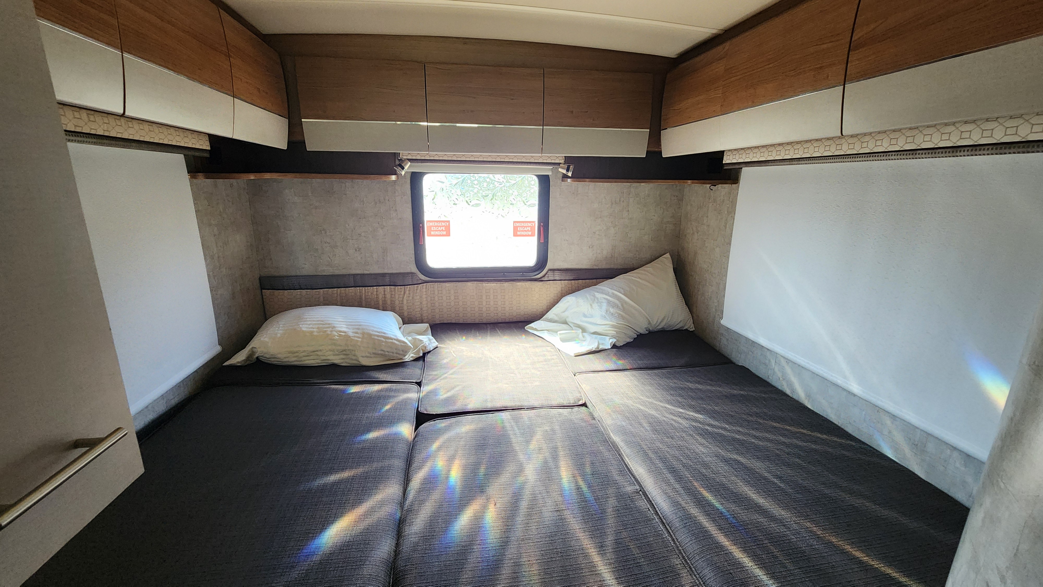 A bright RV rental interior featuring a modular bed with pillows, sleek overhead cabinets, and sunbeams creating rainbows.. Winnebago View 2015