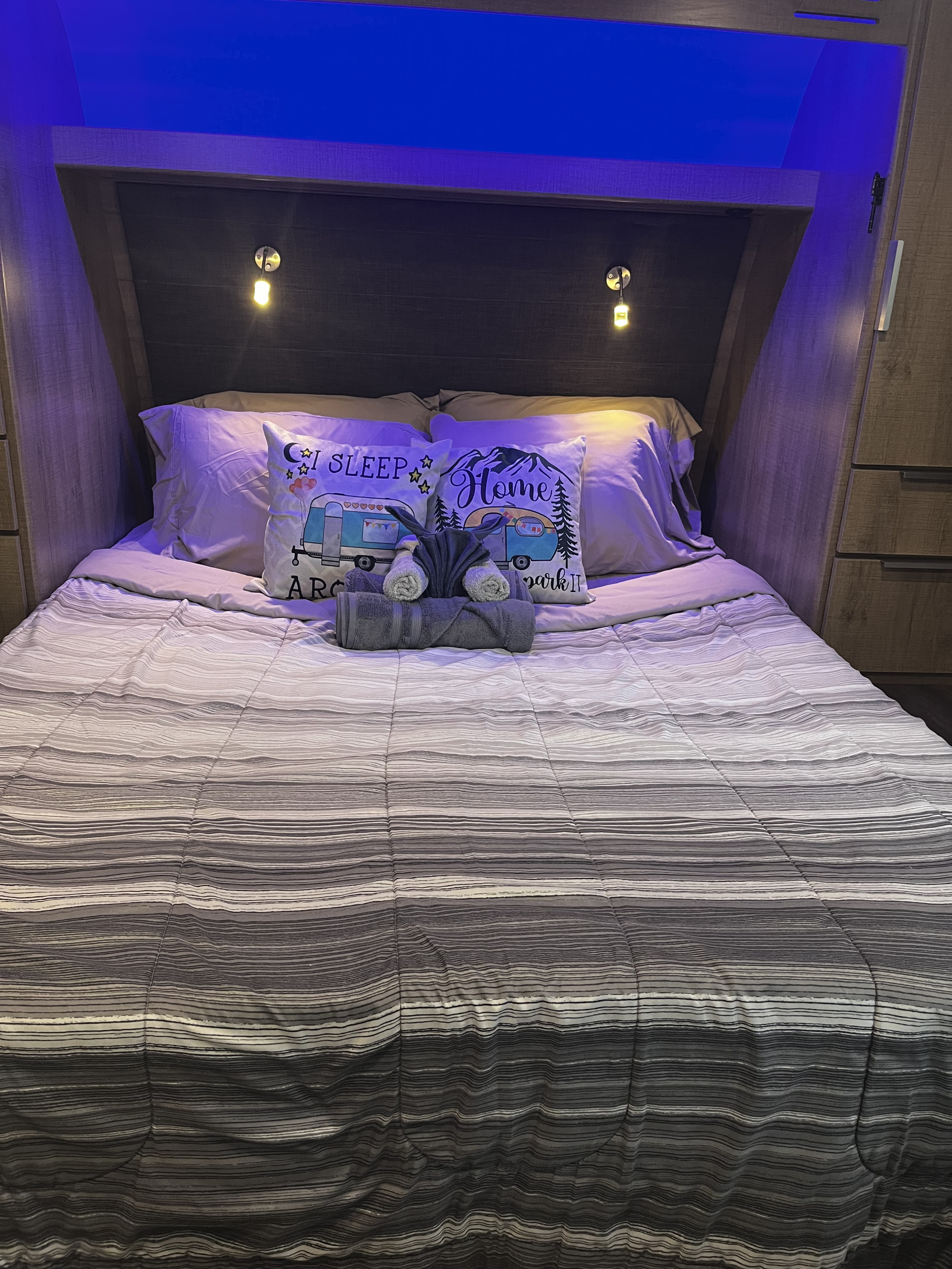 Cozy RV rental bedroom with a striped comforter, "camping" pillows, rolled towels, and blue ambient lighting, perfect for travel.. Grand Design Imagine XLS 2021