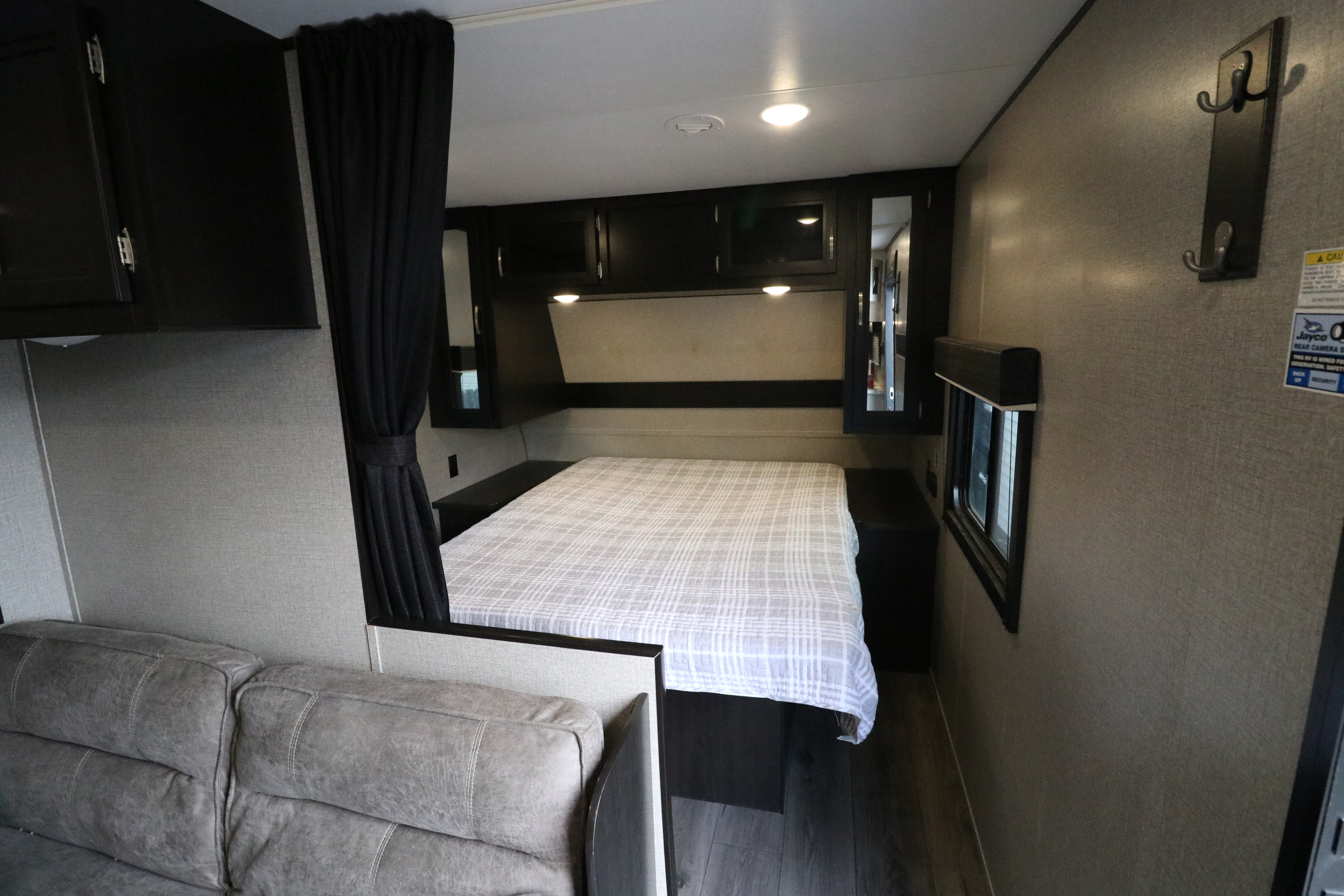 RV rental interior: Features a neatly made bed with plaid spread, dark cabinetry, sofa, and window. A cozy space for travel.. Jayco Jay Feather 2020