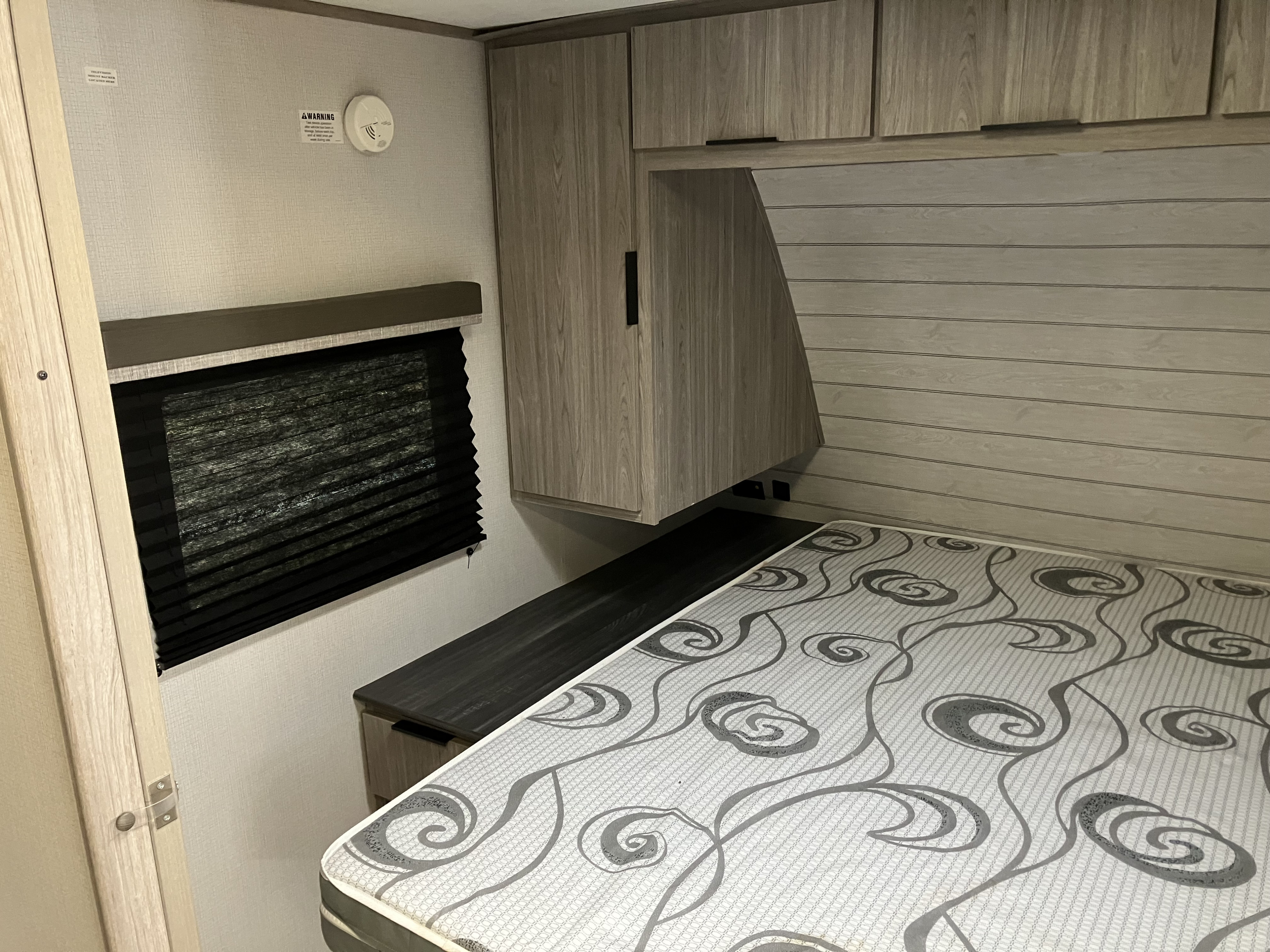 Inside an RV rental bedroom featuring a patterned bed, wood-grain cabinets, window with blind, and a smoke detector.. Keystone RV Passport SL 2021