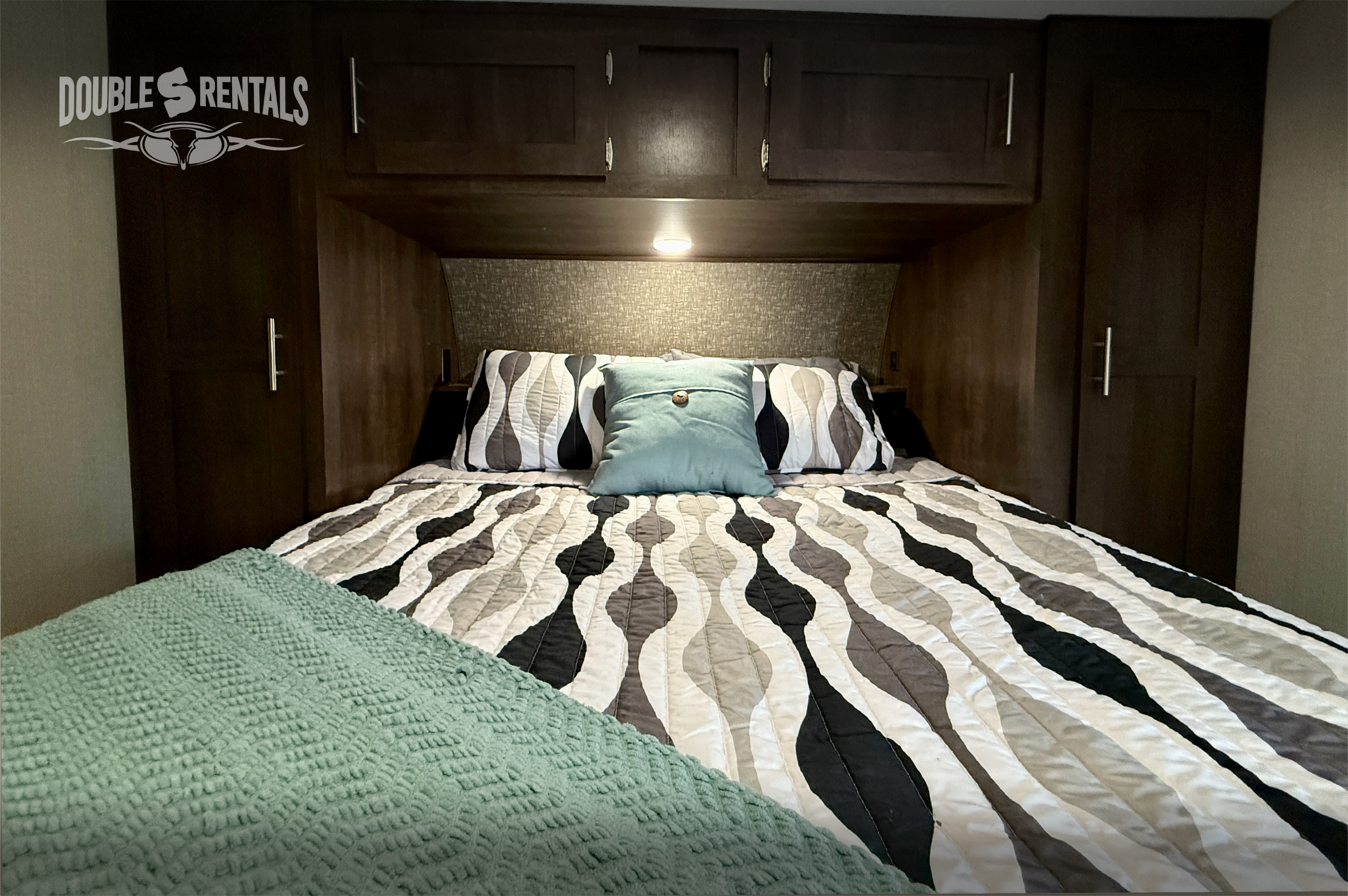 Inside a comfortable RV rental bedroom, featuring a bed with patterned quilt, accent pillows, and dark wood overhead cabinets.. KZ Sportster 2021