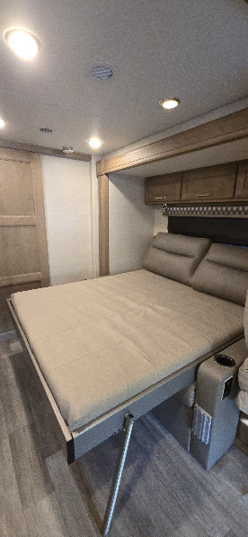 Modern RV rental interior featuring a beige convertible bed with headrests, overhead cabinets, and wood-look floor.. Tiffin Wayfarer 2023