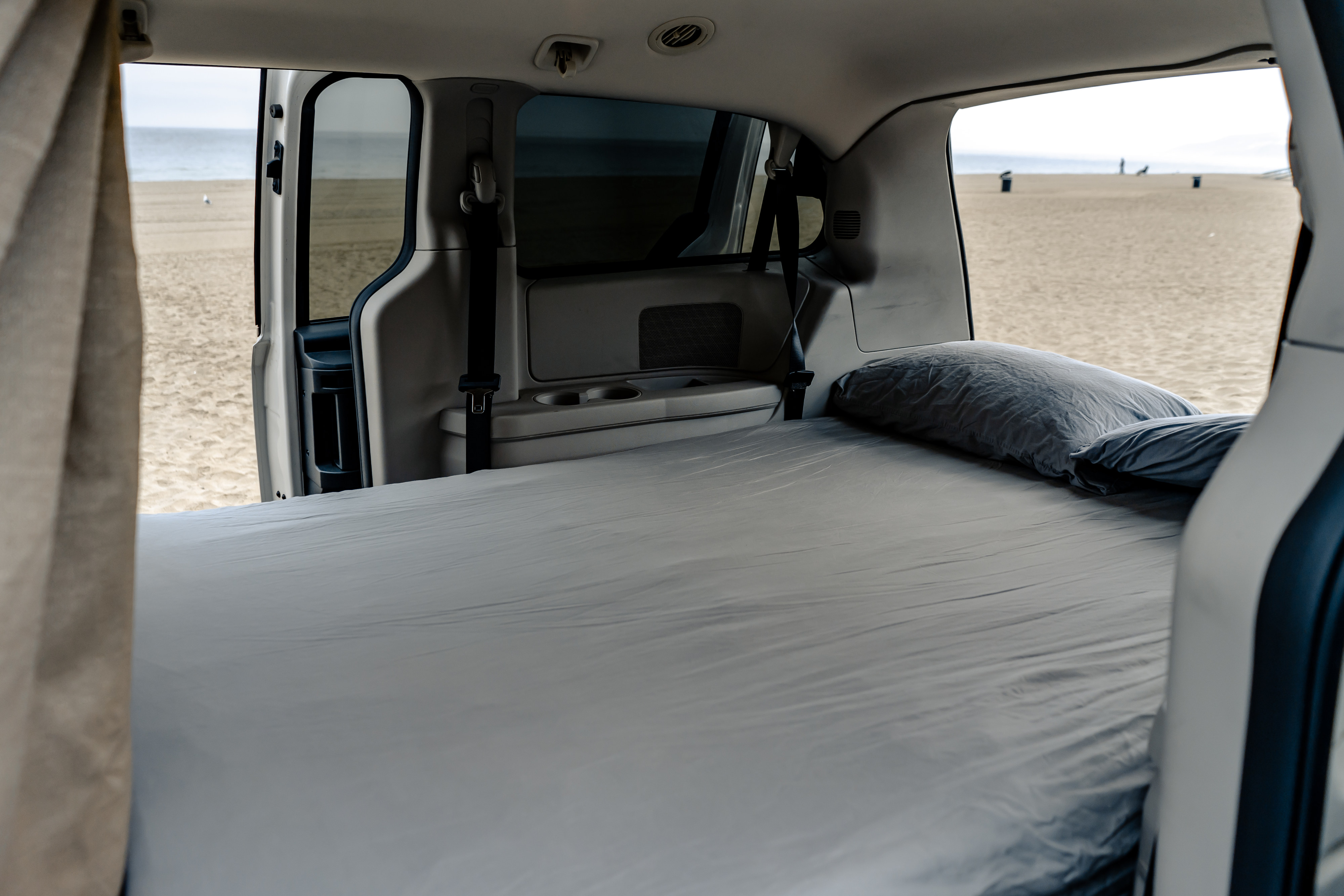 Interior of an **RV rental** showing a bed with gray sheets looking out onto a sandy beach and ocean.. Dodge Caravan 2019