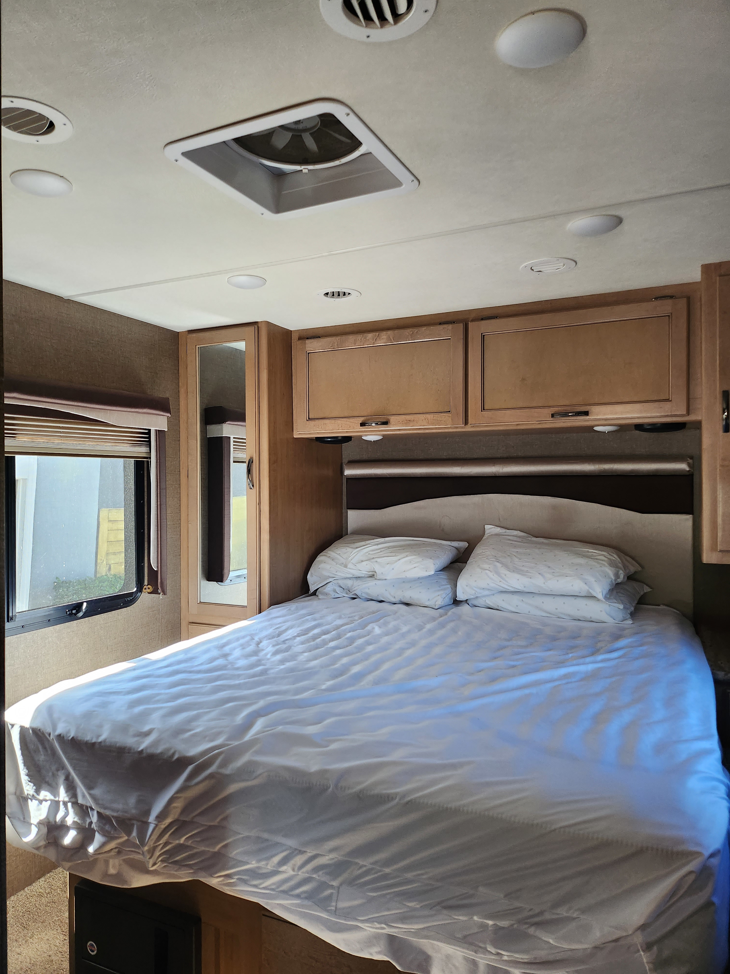 Inside an RV rental, a cozy bedroom with a neatly made bed, white linens, wood cabinets, and a window for natural light.. Thor A.C.E. 2018