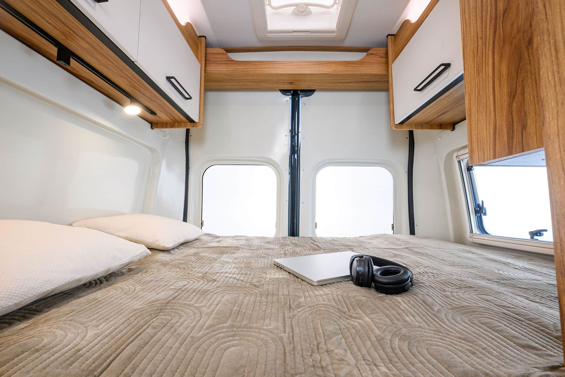 Modern RV rental bedroom: comfy bed, laptop, headphones, wood cabinets, and bright rear windows.. Font Vendome Terravan 2025