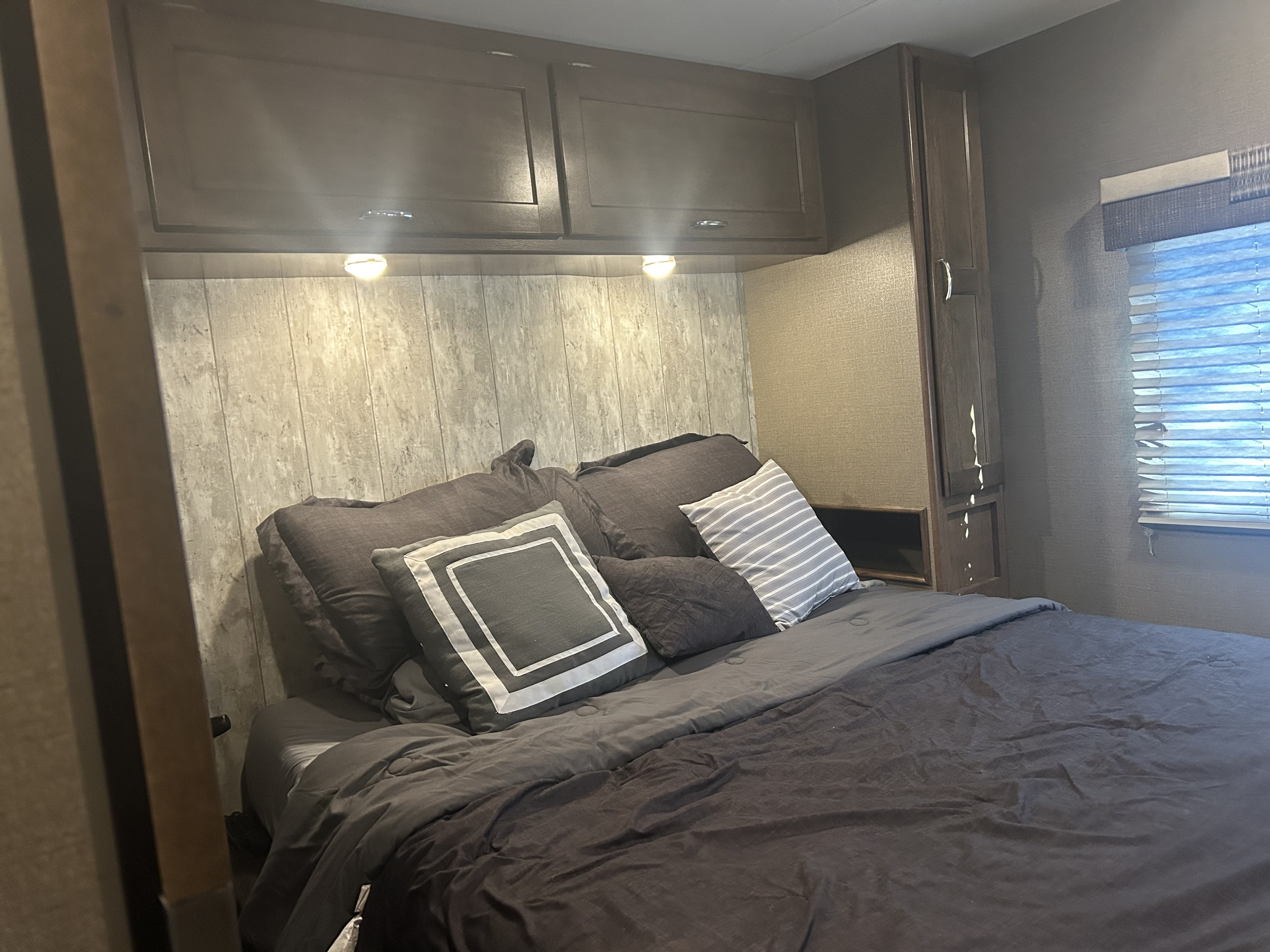 Comfortable RV rental bedroom featuring a bed, wood-paneled wall, overhead cabinets, and window. Ideal for trips.. Winnebago Intent 2018