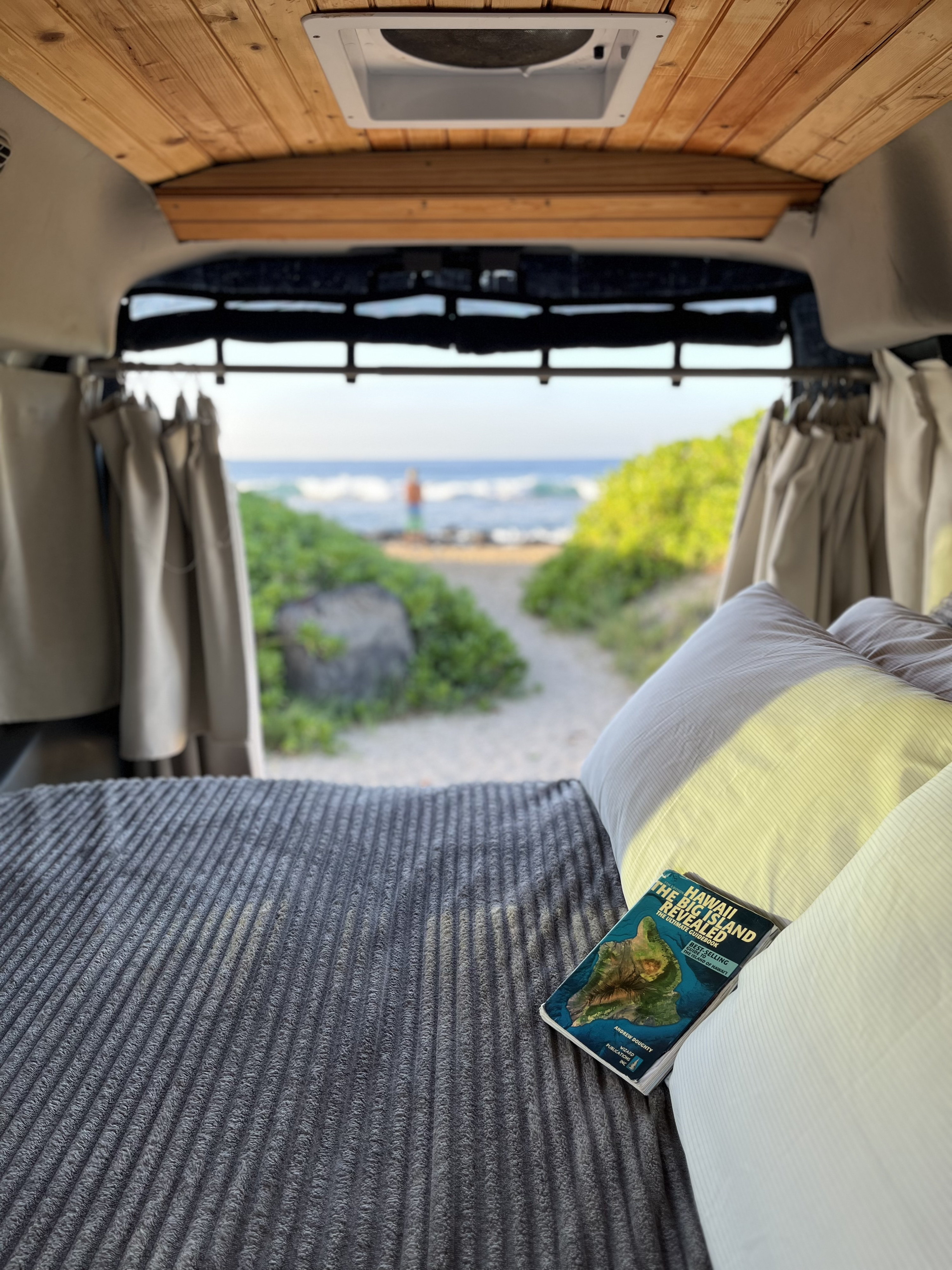 Relaxing inside an RV rental. A Hawaii guidebook rests on the bed, with a serene beach view beyond.. Ford Transit 2015