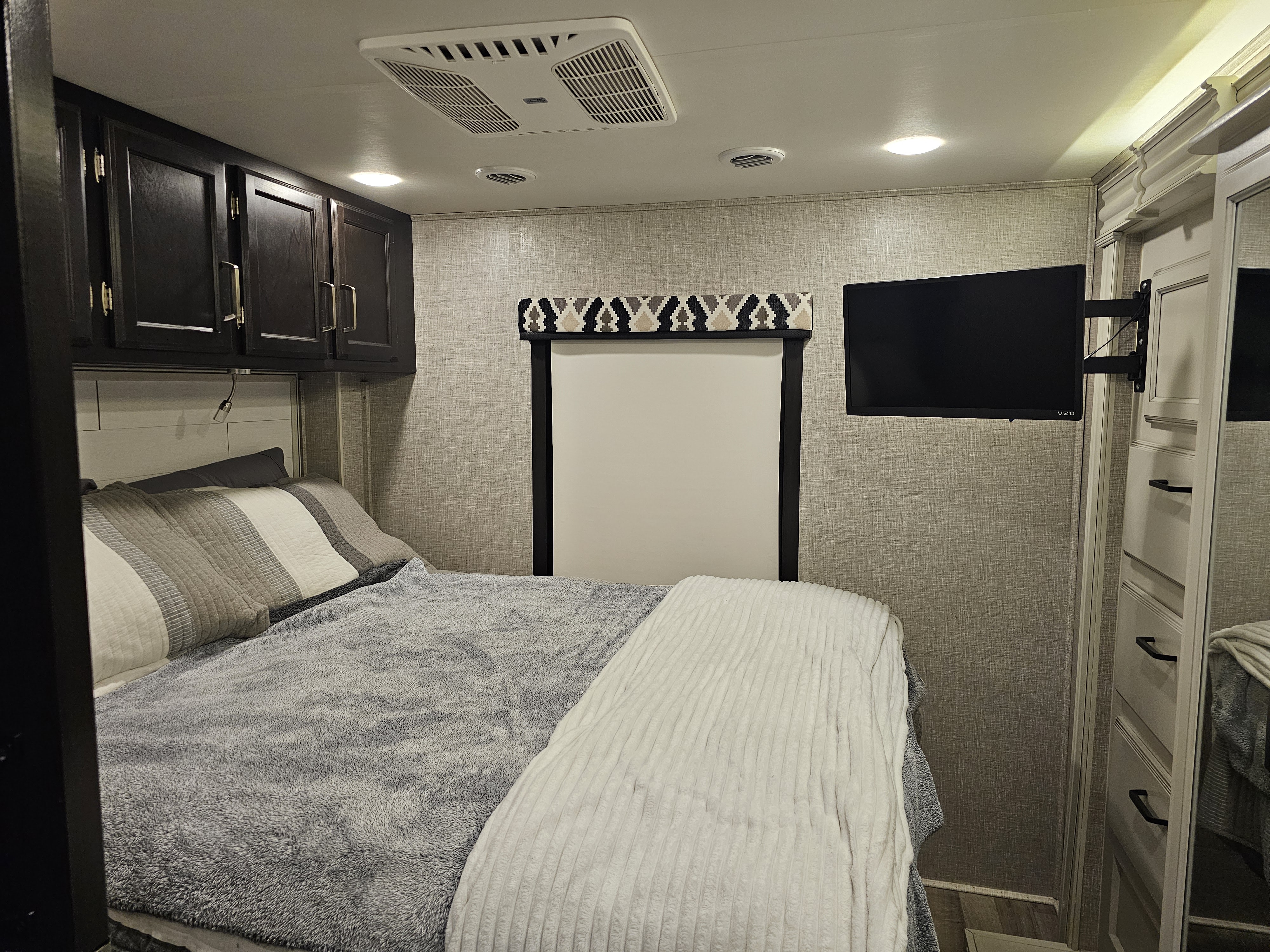 Cozy RV rental bedroom with dark wood cabinets, a wall-mounted TV, and comfortable grey/white bedding.. Jayco Alante 2022