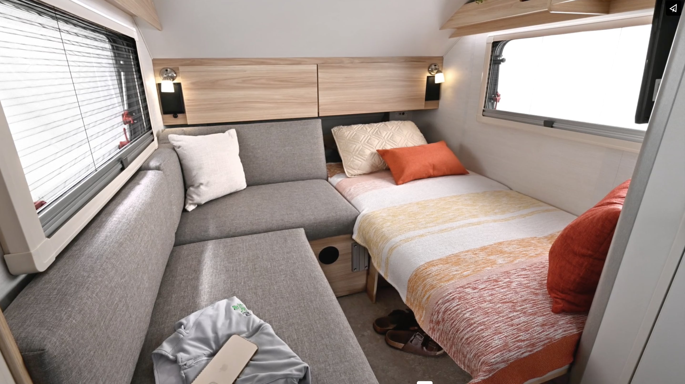 Cozy RV rental interior with an L-shaped gray sofa, striped bed, overhead cabinets, and bright windows.. nuCamp T@B 2024