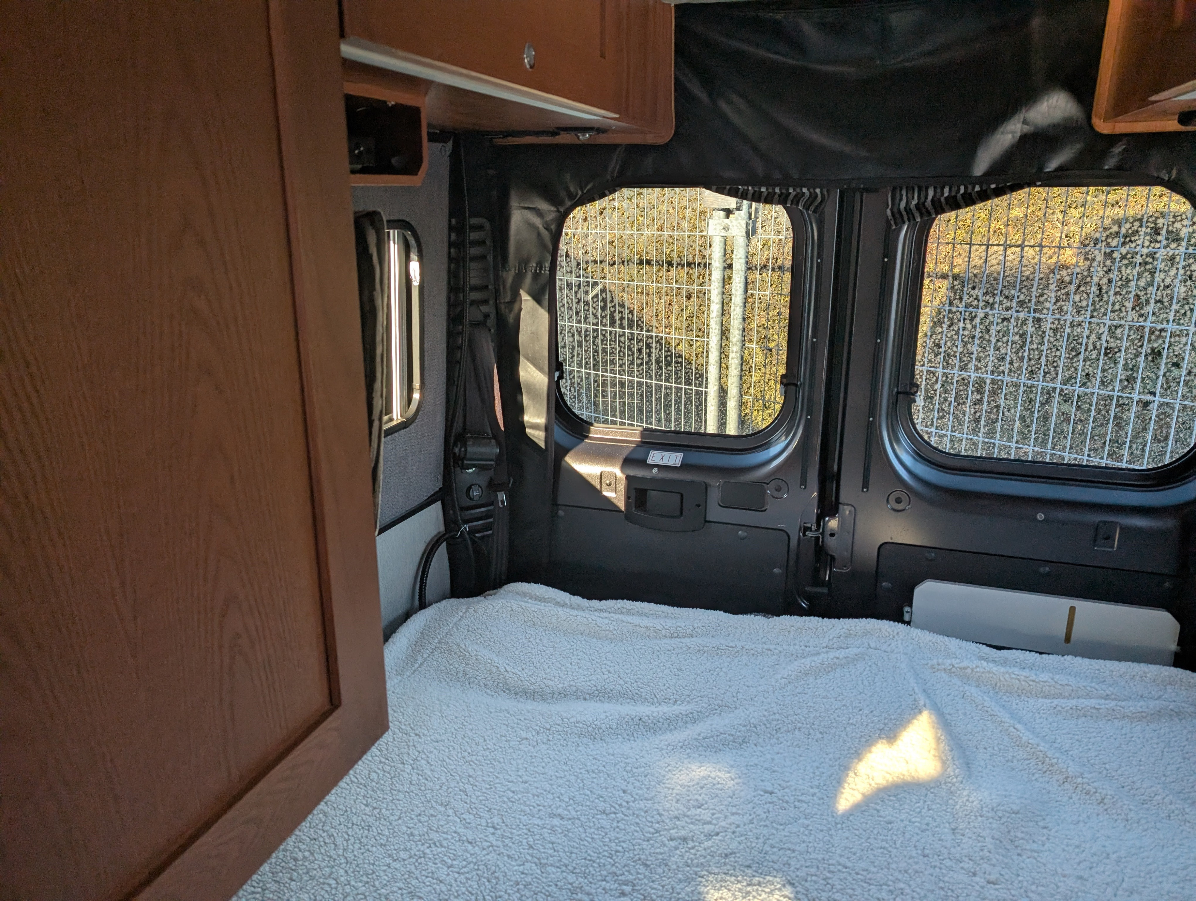 Cozy rv rental interior with a white bed, woody cabinets, and rear windows showing an outdoor fence.. Roadtrek Roadtrek Class B 2019
