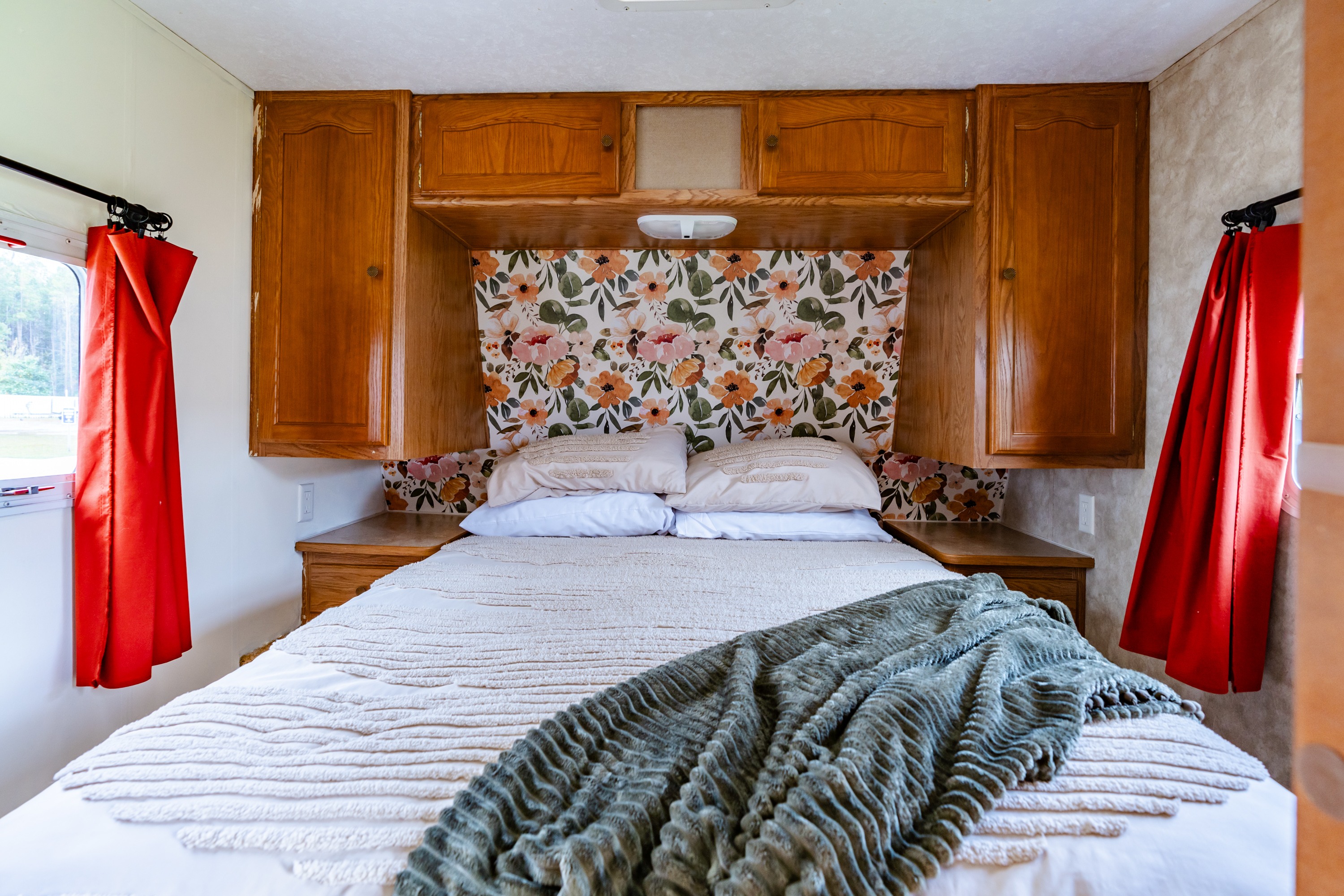 Cozy RV rental bedroom featuring a floral headboard, wood cabinets, and red curtains by the bed.. Keystone RV Springdale 2006