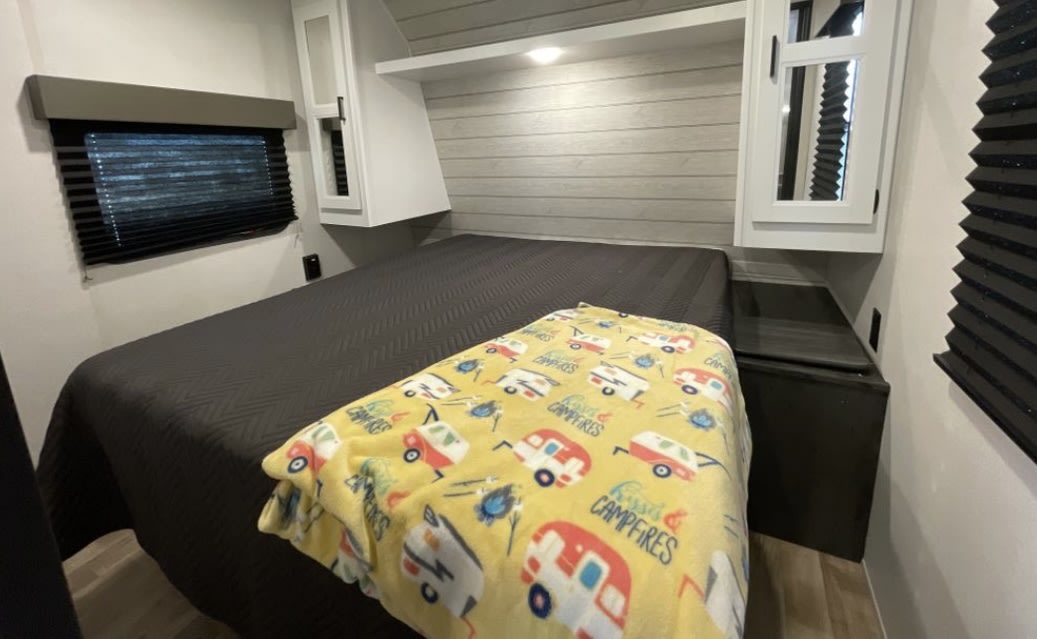 RV rental bedroom features a cozy bed with fun camper blanket, overhead cabinets, and a window with blinds. Ideal for travel.. Keystone RV Hideout 2023