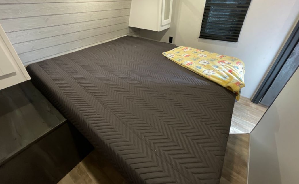 Cozy RV rental bedroom with a dark grey bed, yellow blanket, and light wood interior. Ideal for travel accommodation.. Keystone RV Hideout 2023