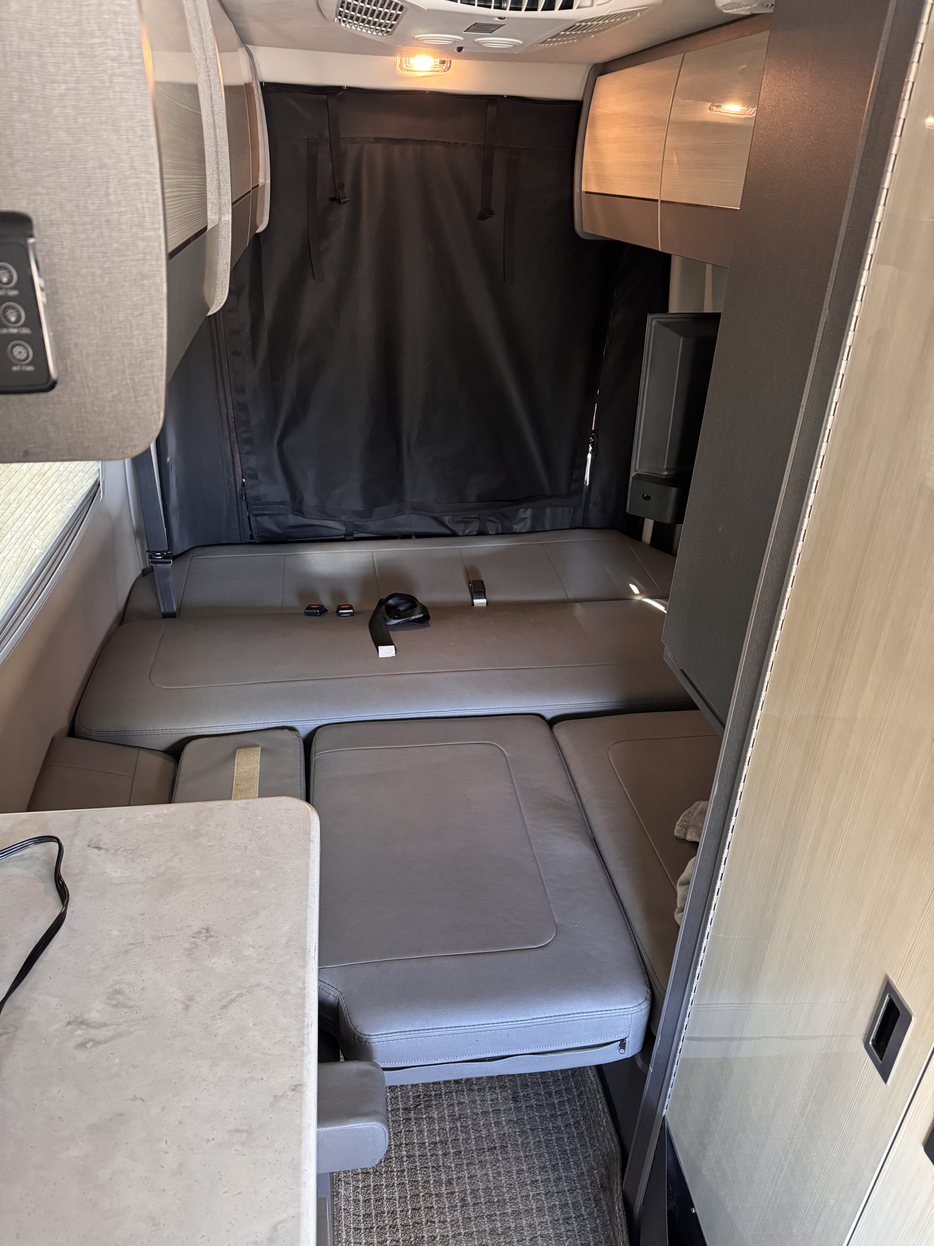 RV rental interior: versatile grey seating converts to a bed, with a black privacy curtain and storage cabinets.. Thor Sanctuary 2024