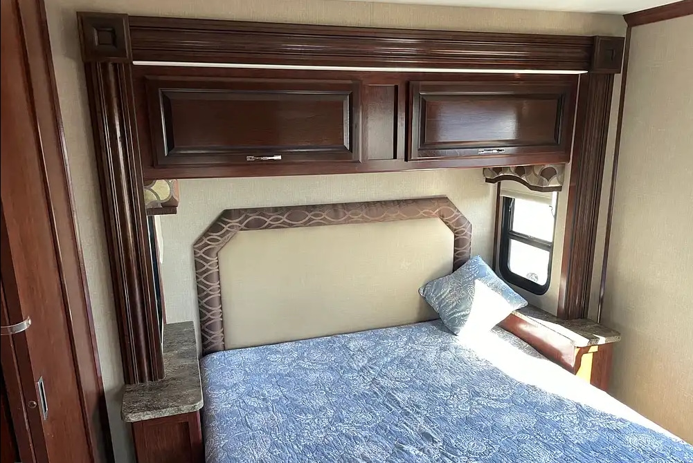 Comfortable RV rental bedroom featuring dark wood overhead cabinets, an upholstered headboard, and a blue patterned bed.. Flair-Fleetwood LXE 2018
