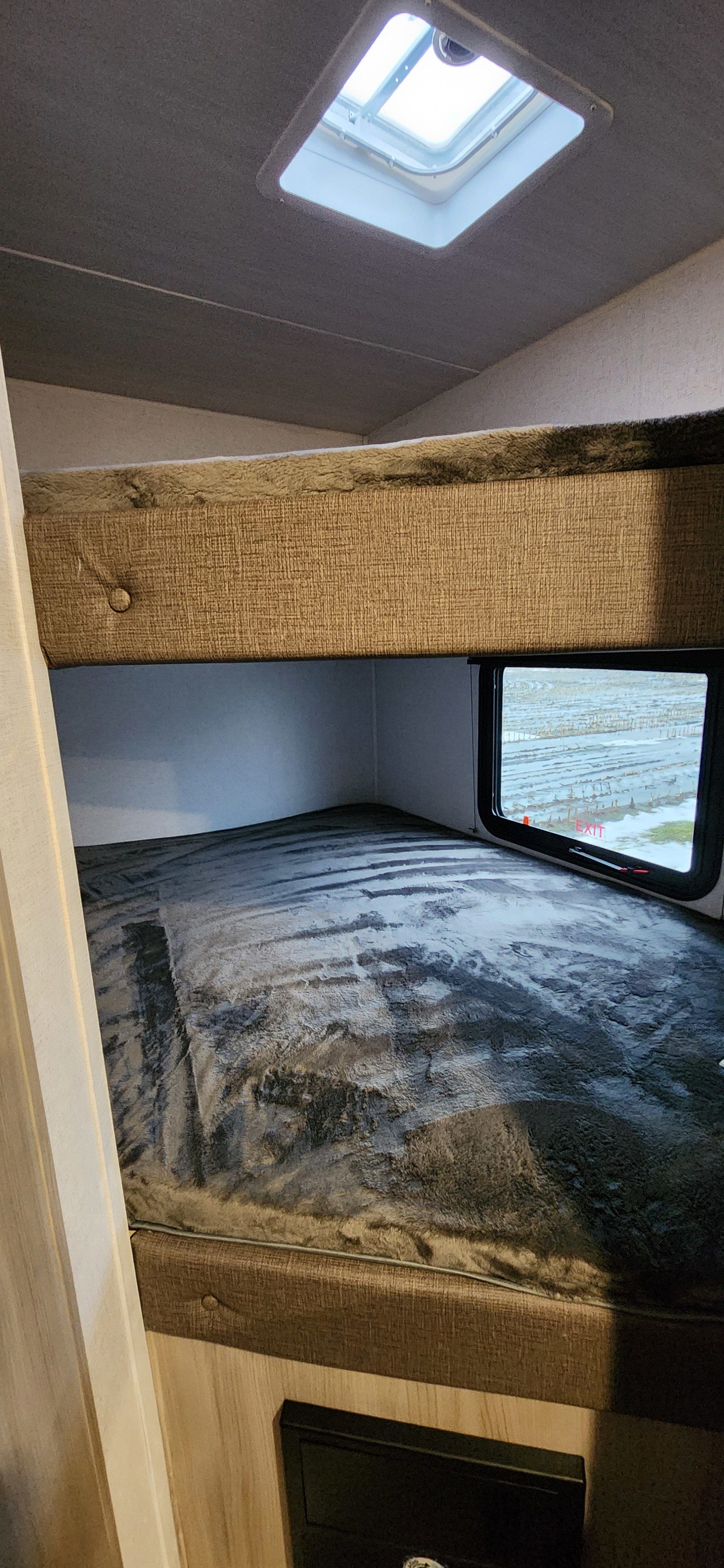 Cozy RV rental bunk area: lower bed with grey blanket, upper bunk above, window, and ceiling vent visible.. East to West Silver Lake 2024