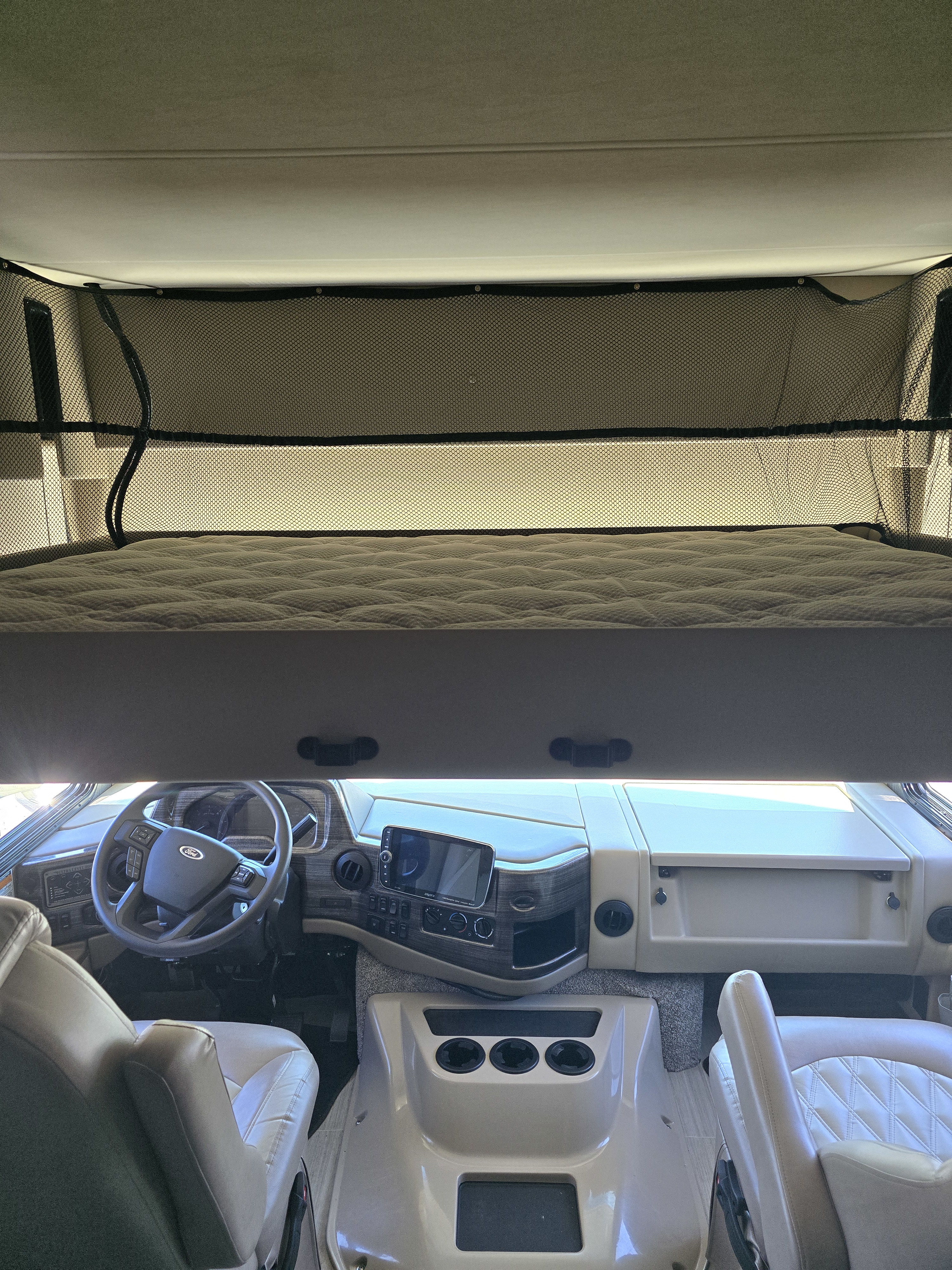 Interior of an RV rental: driver's cockpit with dashboard, steering wheel, and an overhead bunk bed with safety net.. Thor Challenger 2021