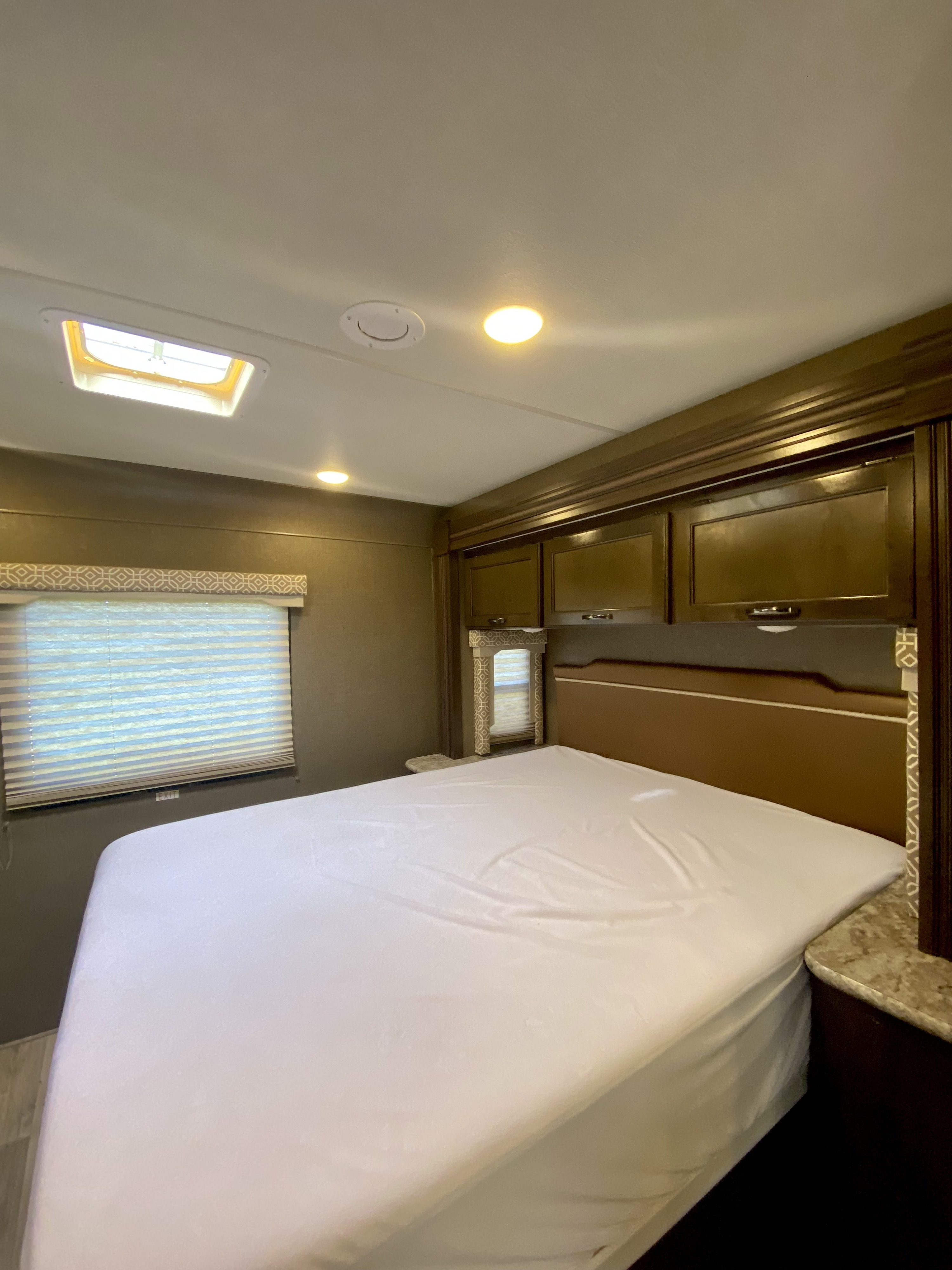 RV rental bedroom: a clean, white-sheeted bed, window with blinds, and dark wood overhead cabinets.. Thor Freedom Elite 2018