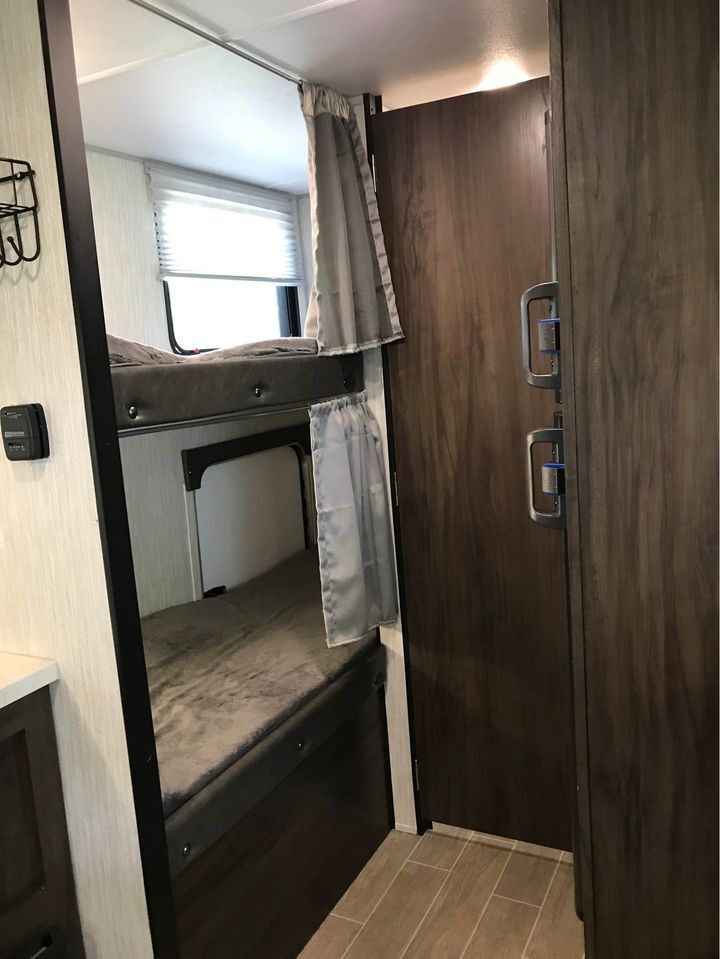 Inside this rv rental, bunk beds with gray bedding and privacy curtains are on the left. A window and a dark wood door are visible.. Forest River Cherokee Wolf Pup 2020