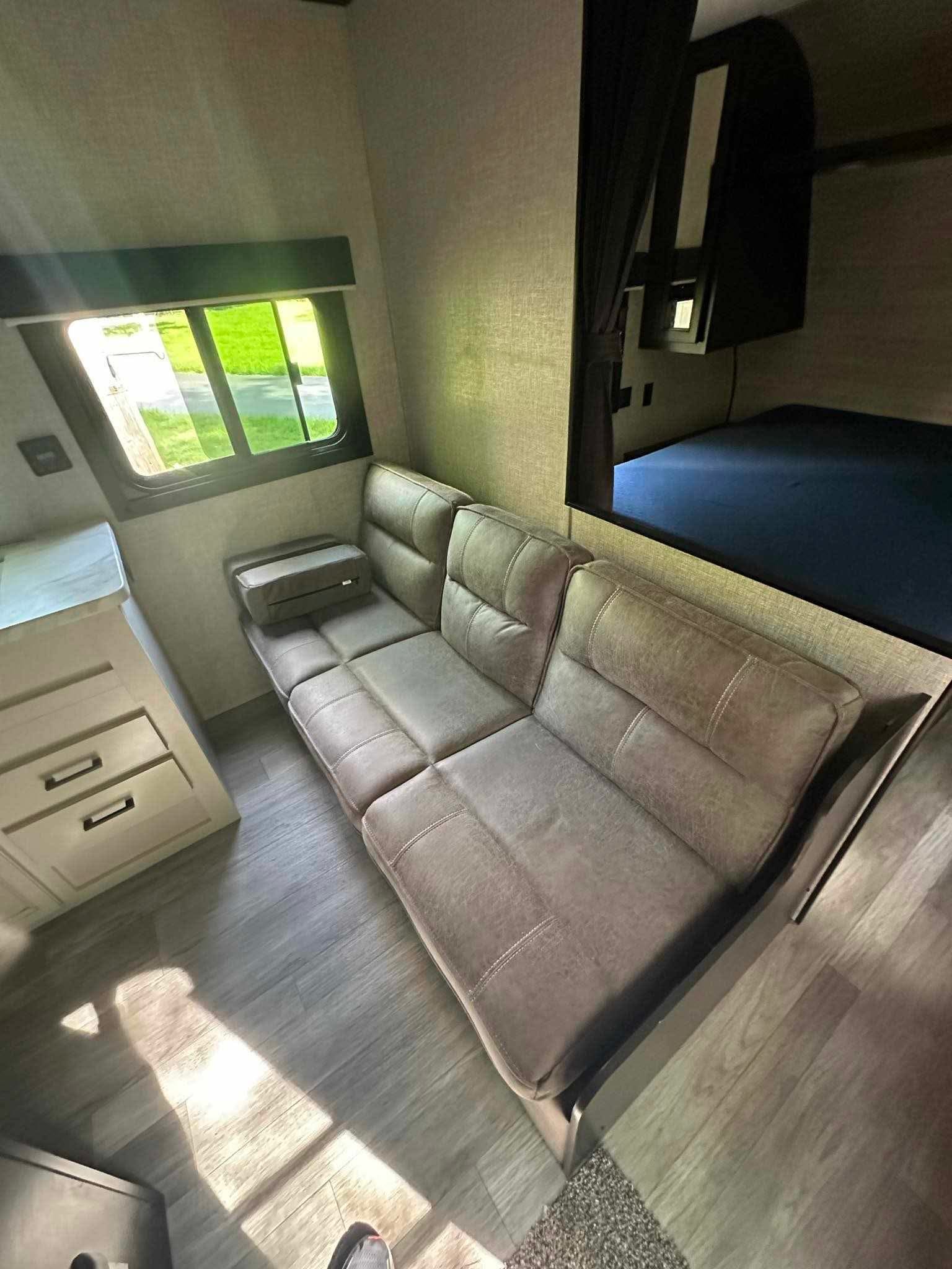 Bright RV rental interior. Features a long sectional sofa, cabinet with drawers, and an upper bunk bed with privacy curtain.. Jayco Jay Flight SLX 2022