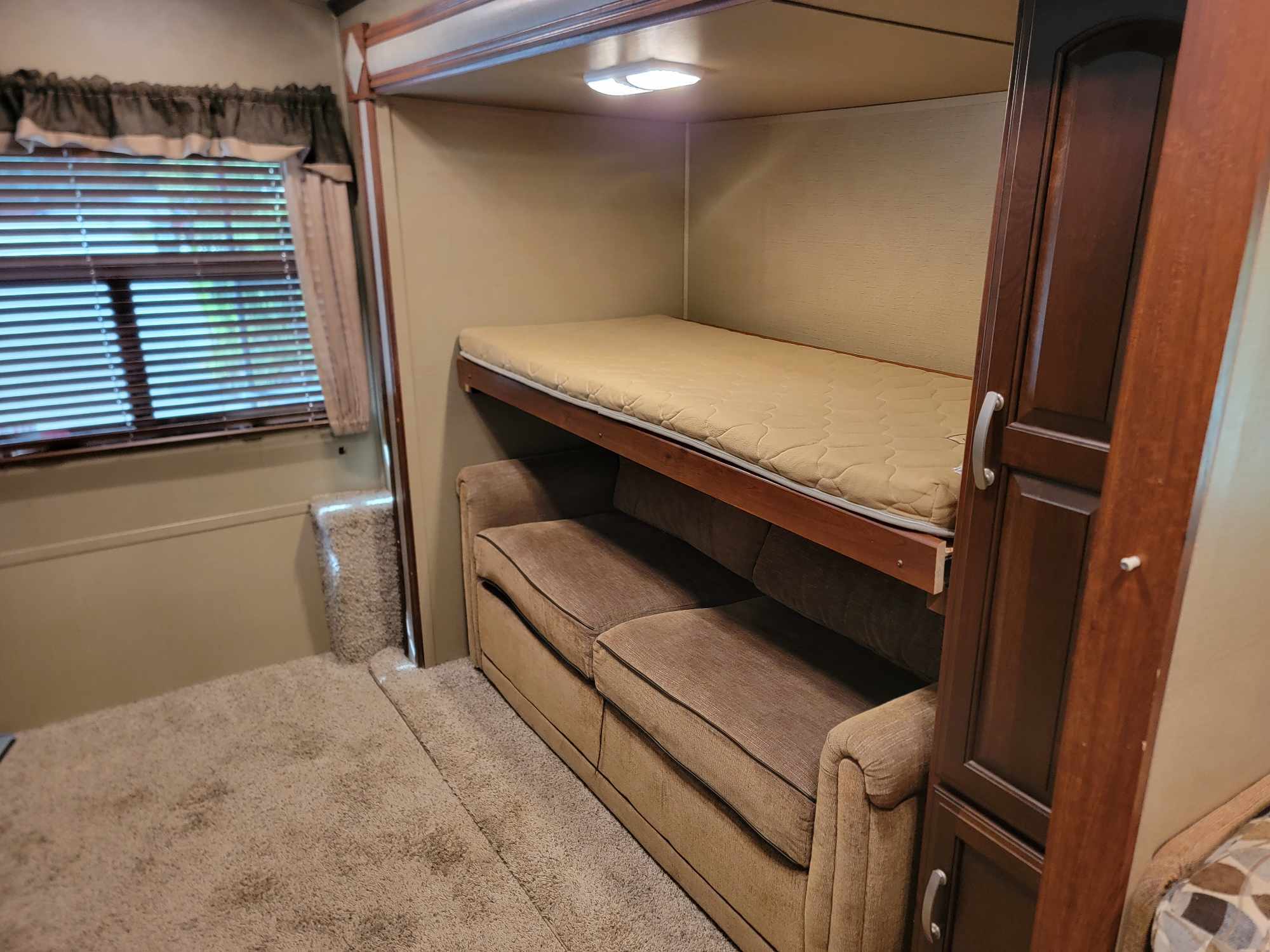 RV rental interior featuring a cozy bunk bed over a sofa, a window with blinds, and storage cabinetry. Ideal for travel comfort.. Keystone RV Outback 2014
