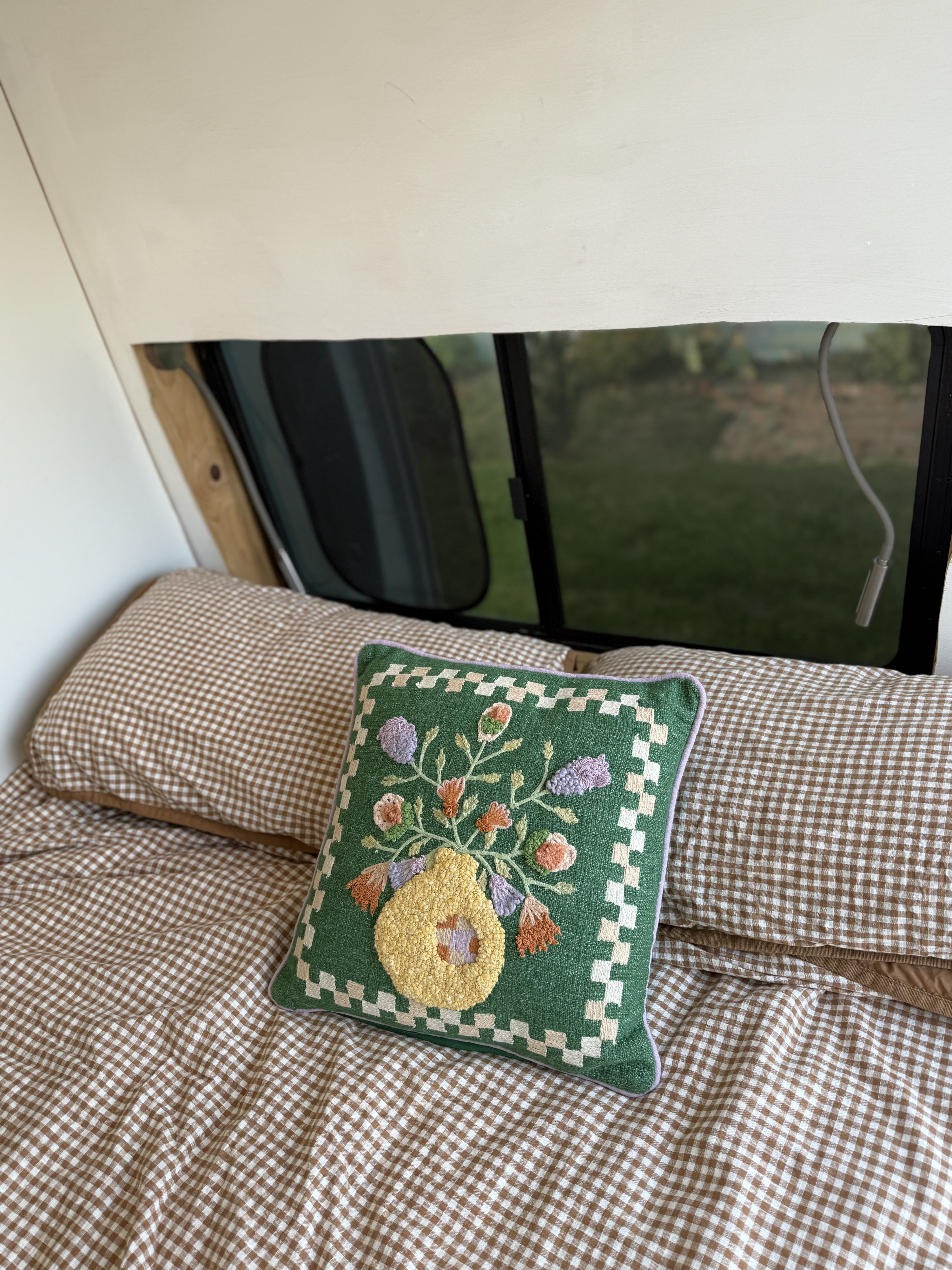 Cozy RV rental interior with brown gingham bedding and a green, floral embroidered pillow by the window.. Ford Transit 2021