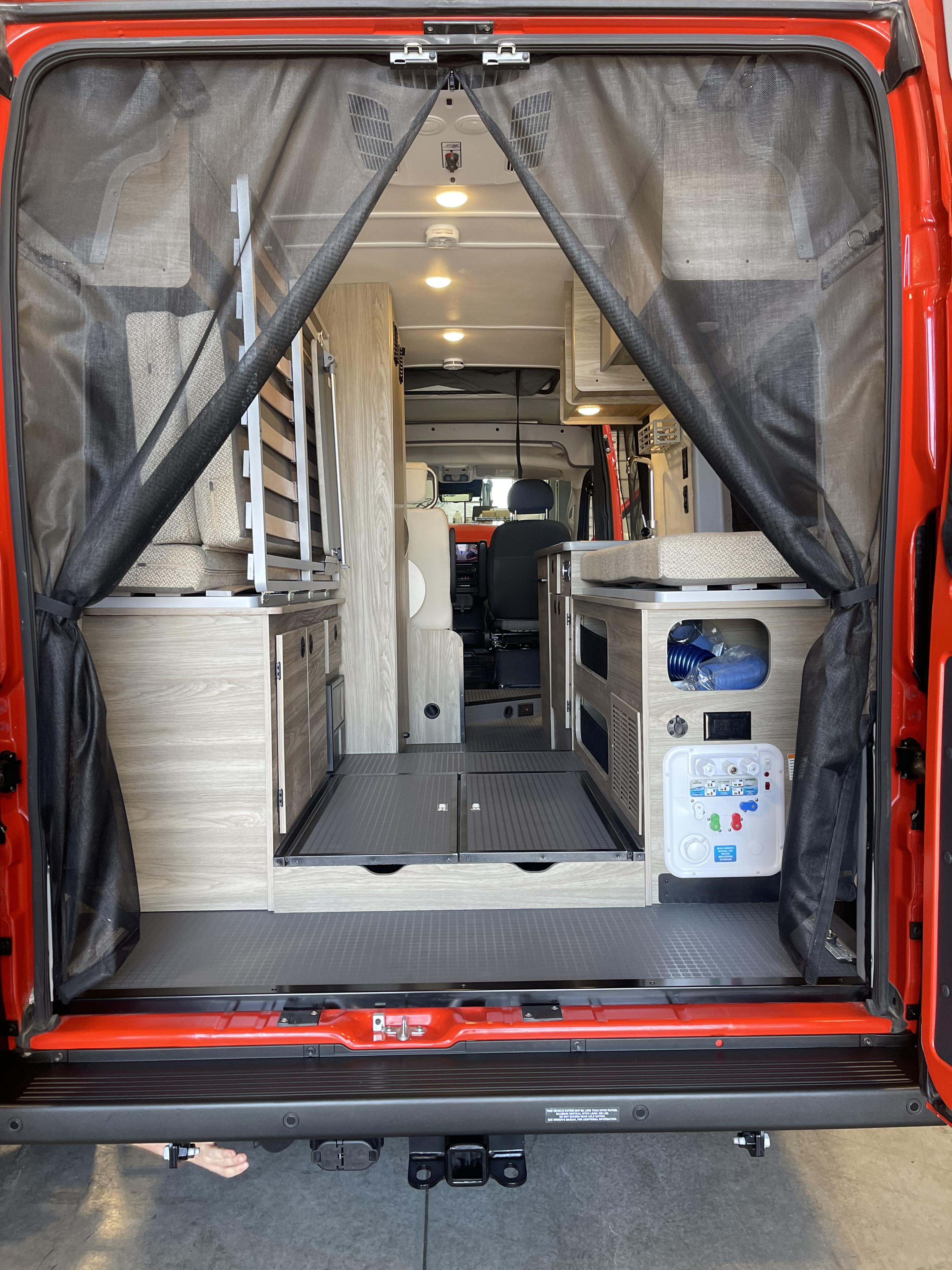 Spacious RV rental interior from the rear of a red camper van, showing a modular bed, kitchenette, and utility panel through mesh screens.. Winnebago Solis 2024