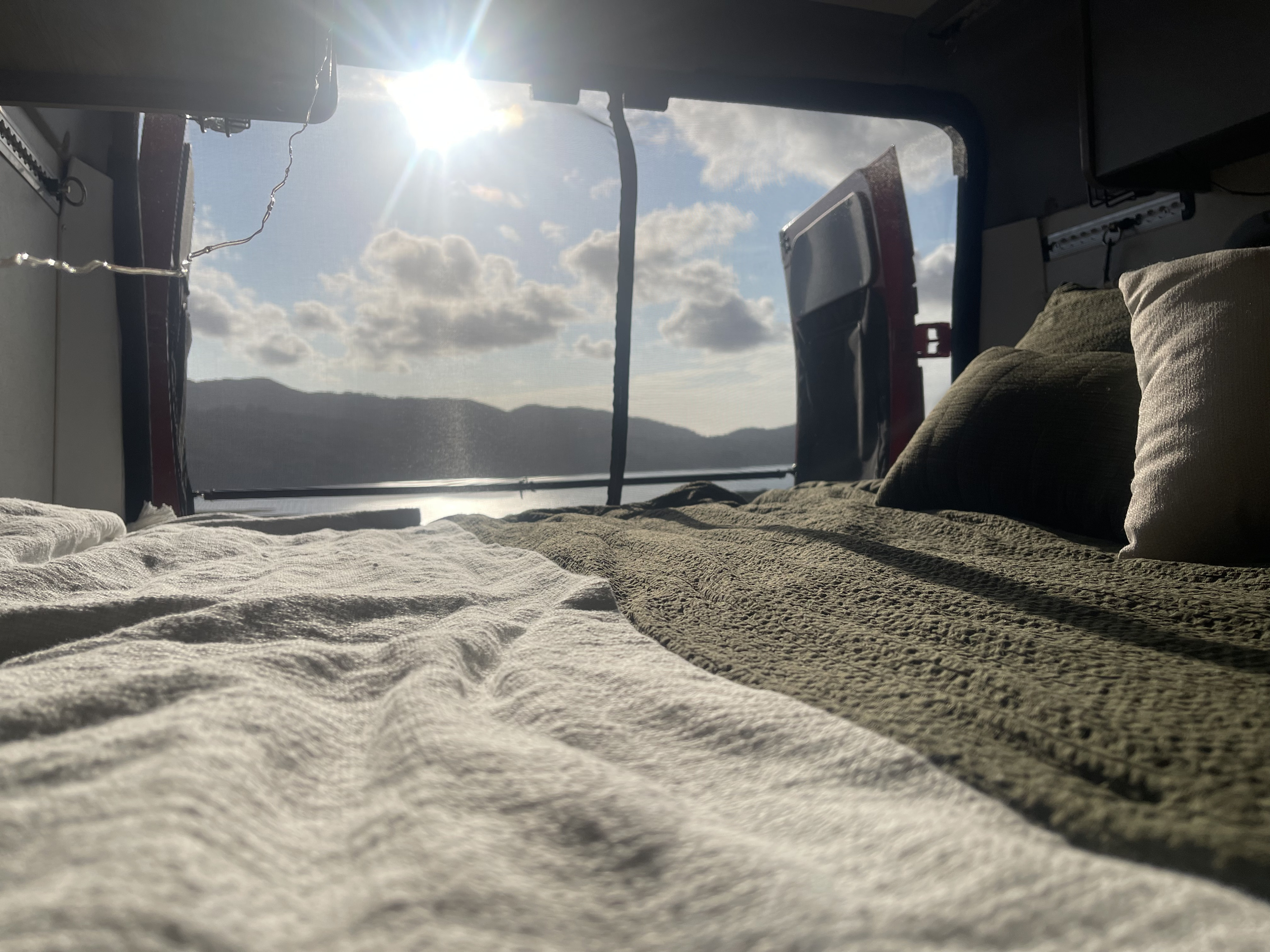 An inviting bed in an rv rental, showcasing a beautiful serene lake and mountain view through the open rear doors on a sunny day.. Winnebago Solis 2024