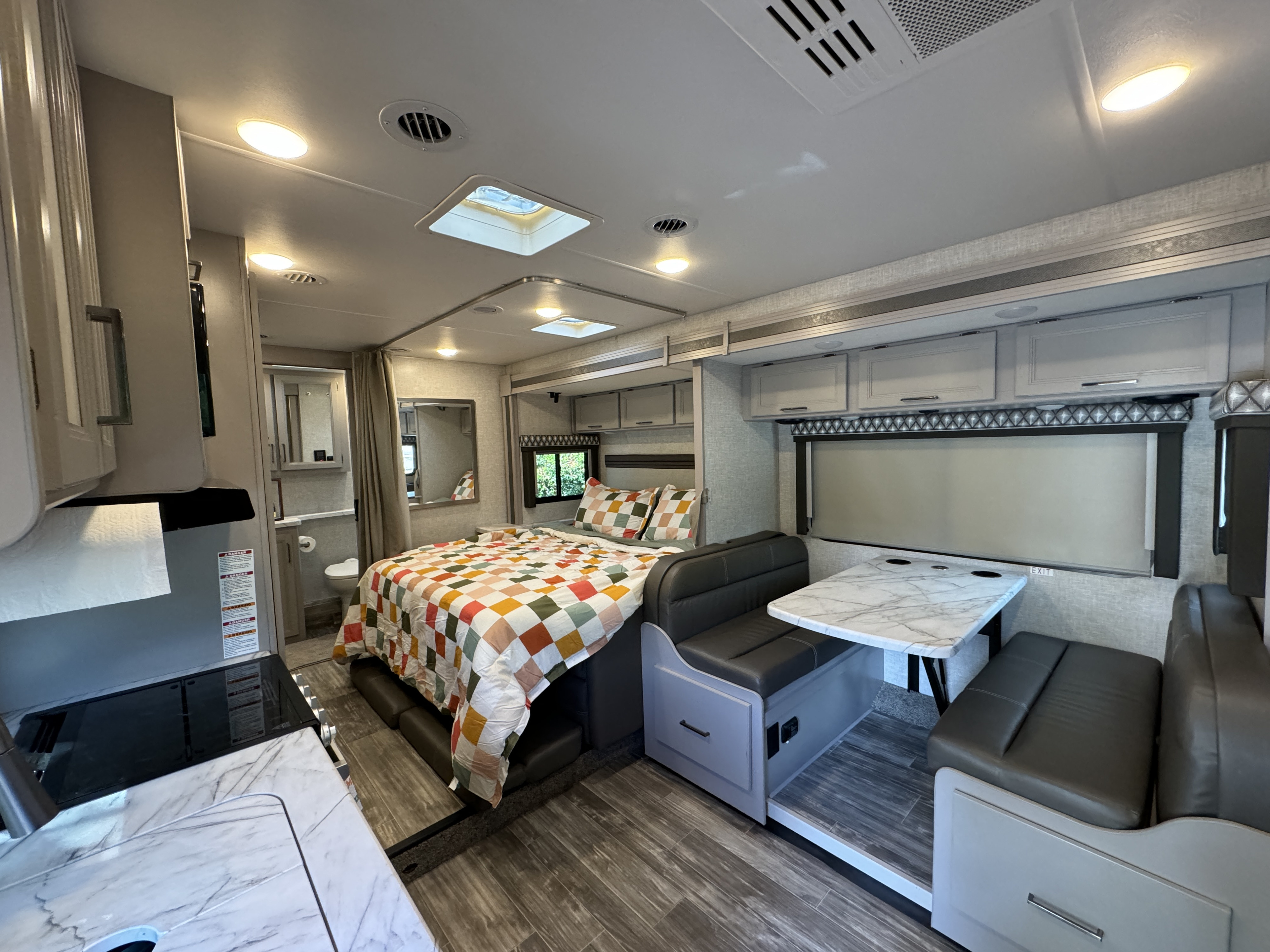 Bright RV rental interior showing a cozy bedroom with colorful quilt, kitchen area, and dining booth with a marble table.. Thor Geneva 2023