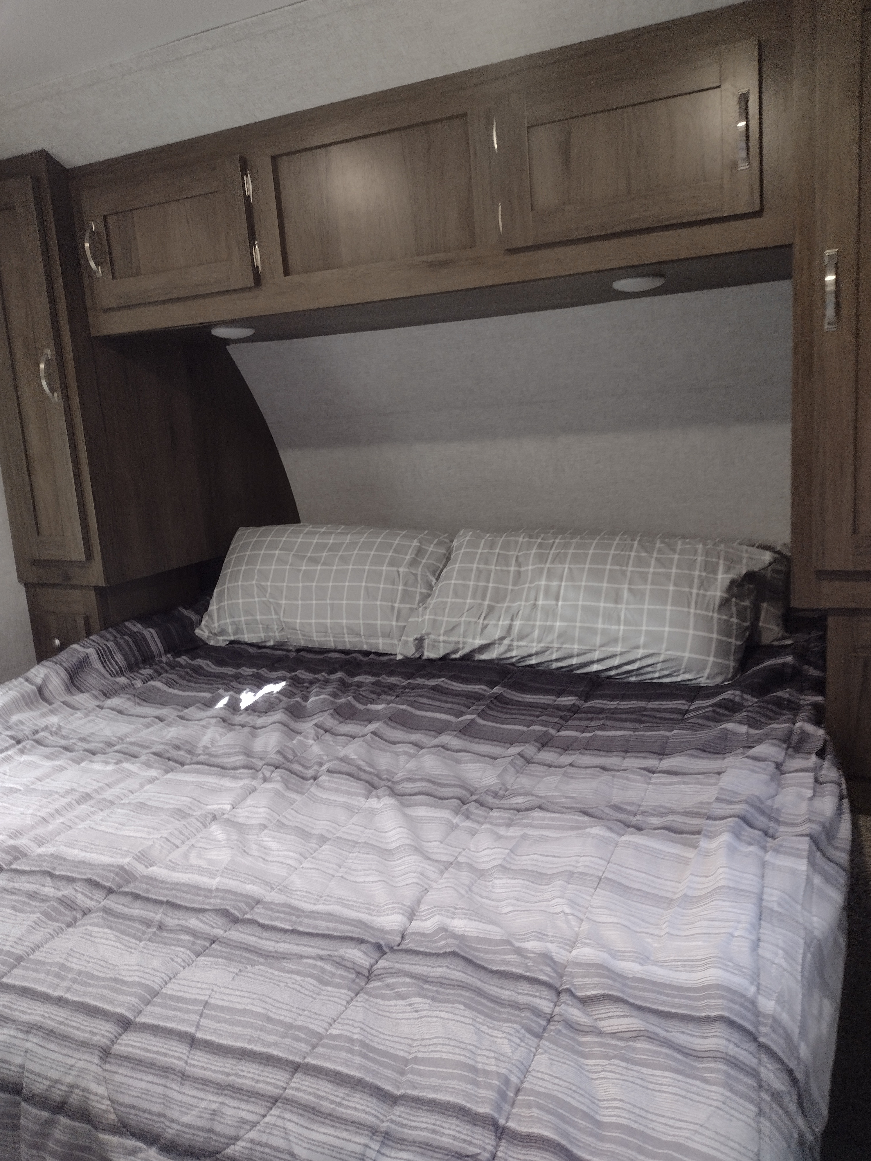 Experience comfort in this RV rental bedroom, featuring a grey striped bed, checkered pillows, and functional overhead cabinets.. East to West Silver Lake 2021