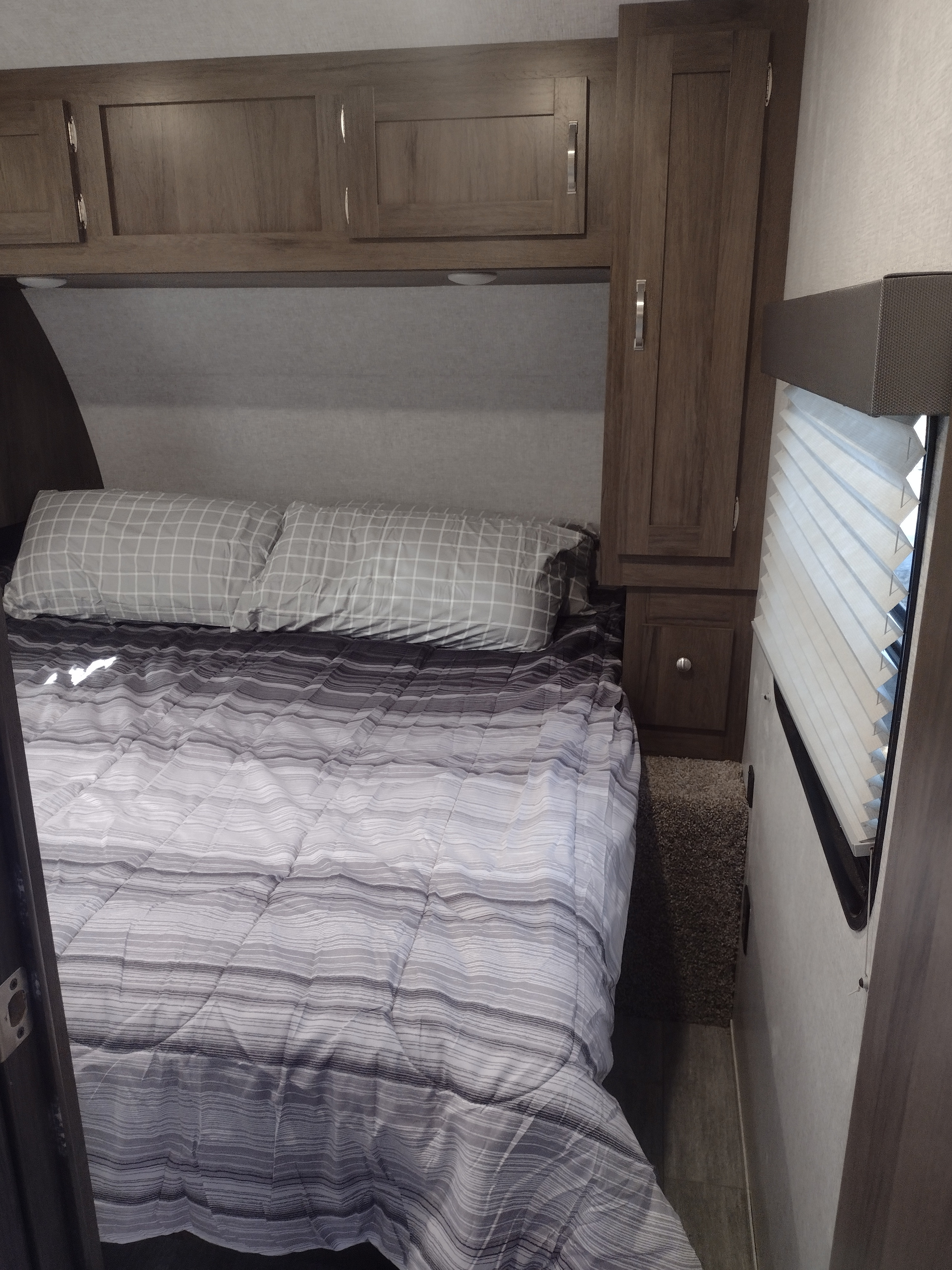 Cozy rv rental bedroom features a comfy bed with striped comforter & checkered pillows, storage cabinets, & window blinds.. East to West Silver Lake 2021